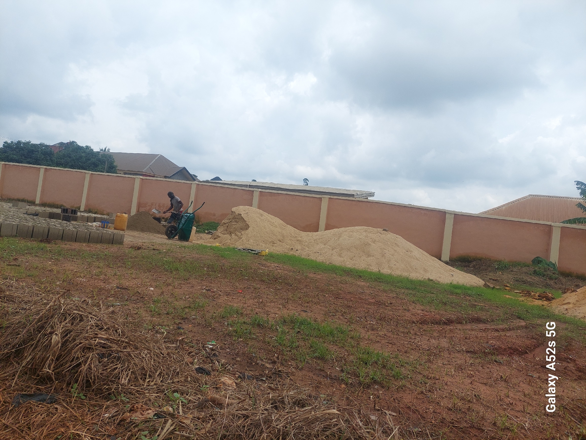 A low-angle, vertical, medium shot captures a scene in Umudioka, Nigeria, with a construction or landscaping area. A pile of light brown sand dominates the foreground, sloping upwards and to the right. To the left of the sand pile, dry, brown grass and some sparse green vegetation are visible, interspersed with scattered debris. A wheelbarrow, primarily green with black accents, is positioned near the sand pile, containing a small amount of sand. Two figures, appearing as construction workers, are near the wheelbarrow; one is bent over, seemingly interacting with the wheelbarrow, while the other stands upright, facing away from the camera. A light beige building with a cream-colored trim runs along the right side of the frame, its roofline visible at the top edge. In the background, lush green trees and more buildings are partially obscured. The sky is overcast with soft, white clouds. The bottom left corner of the image displays the text "Galaxy A52s 5G."