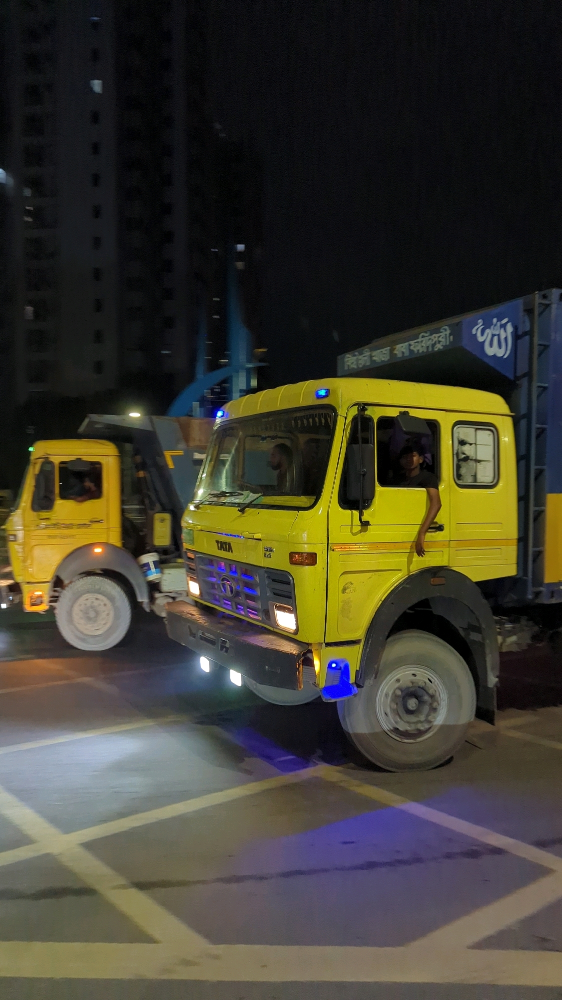 Two yellow Tata trucks are driving on a road at night in Dhaka, Bangladesh. The truck in the foreground is a dump truck, with its bed raised. The truck in the background is a flatbed truck. Both trucks have their headlights on and are illuminated by streetlights. A person is visible in the driver's seat of both trucks. The road has yellow markings on it, indicating a crosswalk. In the background, there are tall buildings with lights on. There is also a blue sign with writing on it, possibly a directional sign or advertisement. The overall scene is dark and appears to be an urban environment.