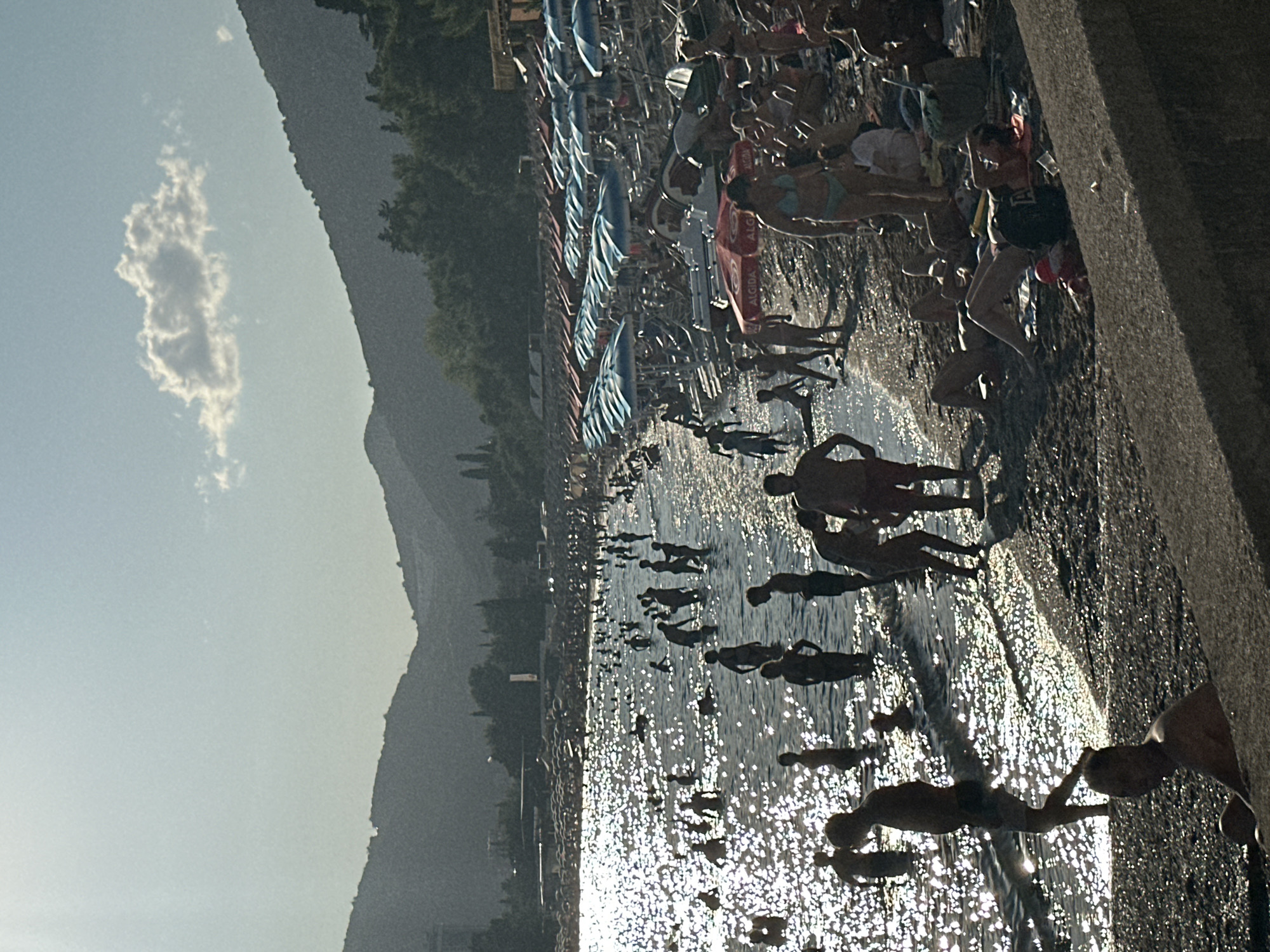 This is a low-angle, wide shot of a crowded beach in Budva, Montenegro, on a sunny day. The scene depicts a busy shoreline with numerous people wading in the water and sunbathing on the beach. The water is sparkling with sunlight, and many individuals are silhouetted against it. In the background, rows of beach umbrellas provide shade, and beyond that, lush green hills rise to form the backdrop, with a clear blue sky overhead. The beach itself is a mix of sand and pebbles. Several people are visible in the foreground, including a man sitting on a wall and others lying on towels. The overall impression is one of a lively summer day at a popular tourist destination.