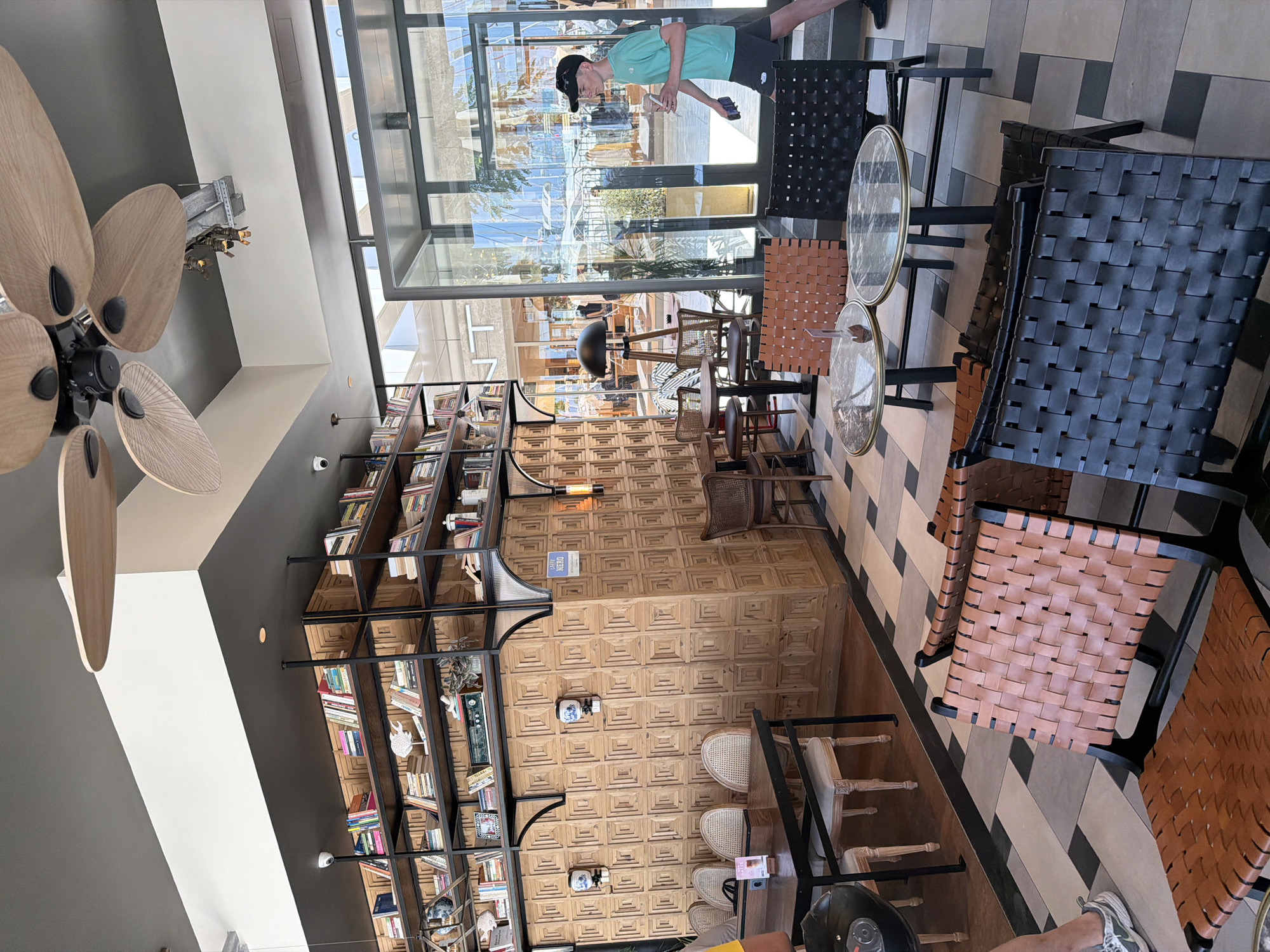 The image depicts the interior of a cafe in Kuşadası, Turkey. A young man wearing a teal t-shirt and black shorts is walking out of the cafe through glass doors. He is carrying a cup in his right hand and a phone in his left. Inside the cafe, there are several tables and chairs, including woven leather chairs in brown and black. A large bookshelf filled with books lines one wall, and decorative wooden panels cover another. A ceiling fan with wooden blades is visible overhead. The floor is tiled in a black and white geometric pattern. The environment appears to be a modern cafe with a mix of industrial and rustic design elements.
