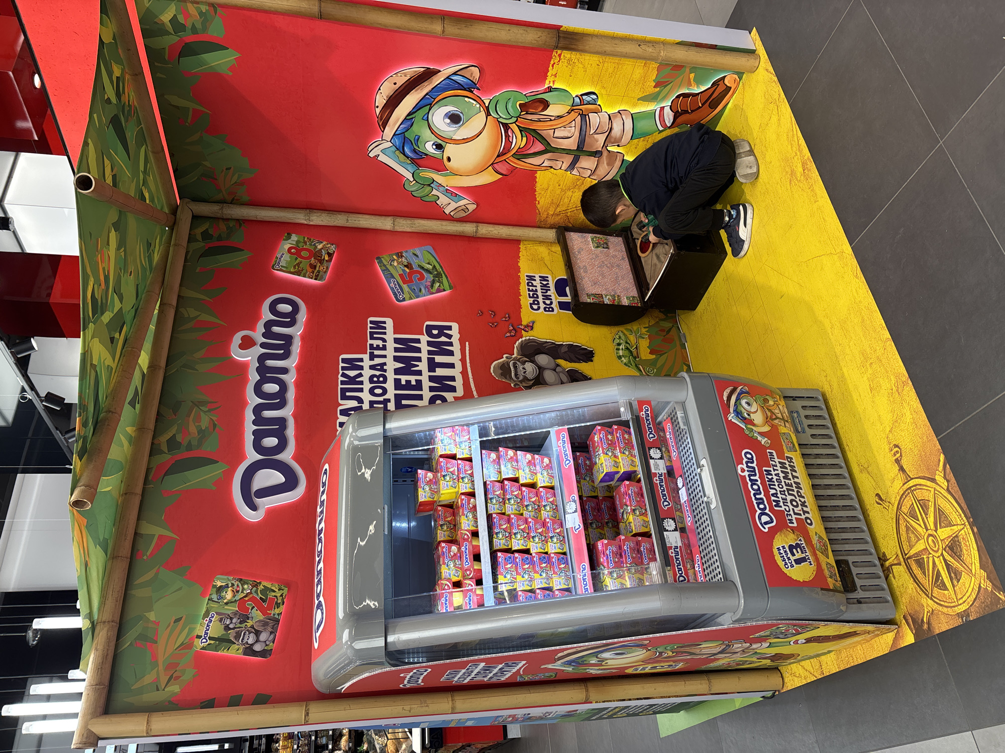 An indoor promotional display for Danonino yogurt is depicted within a supermarket in Sofia, Bulgaria. The display, designed with a jungle or safari theme, features a structure constructed from bamboo-like poles supporting a green, leaf-patterned fabric canopy. The back and side walls are red, adorned with the large "Danonino" logo and Bulgarian text that translates to "SMALL EXPLORERS, GREAT DISCOVERIES." Cartoon illustrations of jungle animals and a safari-clad, dinosaur-like explorer holding a map and magnifying glass are prominent. A gray, glass-fronted refrigeration unit, fully stocked with various colorful Danonino yogurt products, is integrated into the left side of the display. The unit's base includes a sticker promoting "COLLECT ALL 13 MAGNETS." A custom yellow floor mat with a cracked texture and a compass rose design covers the display area, contrasting with the dark gray tiled supermarket floor. A young child, dressed in a dark blue long-sleeved shirt, dark pants, and white Puma sneakers, kneels on the right side of the display. The child is facing and interacting with the contents of an open, dark brown toy chest, which appears to contain small items related to the promotional magnet collection. A light-colored cushion or similar object is positioned under the child's knees. Overhead fluorescent lighting fixtures are visible in the background.