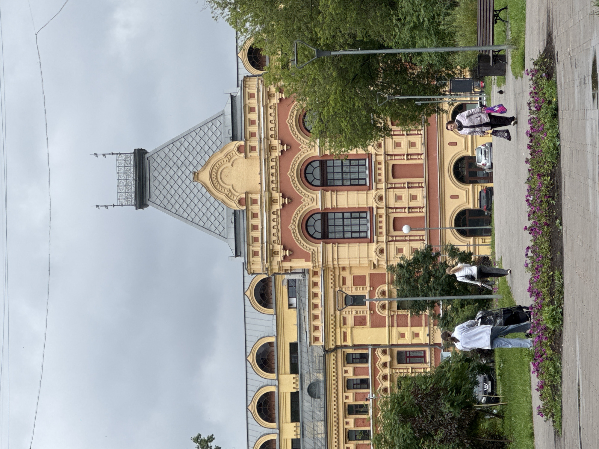 In Nizhny Novgorod, Russia, an overcast day is depicted with a grand, ornate building in the background, featuring a red and yellow facade, arched windows, and a tall, grey mansard roof topped with a decorative spire. In the foreground, a paved walkway meanders through a park-like setting with trees and a flower bed. Two women are visible on the walkway. One woman, in the mid-ground to the left, is pushing a stroller. Another woman, further to the right, walks towards the foreground, carrying two shopping bags. A bench and a trash receptacle are situated to the right of the walkway, adjacent to a dense line of trees. Several cars are parked in the background, near the building's entrance. The overall scene suggests a public space in a city, with pedestrians going about their day.
