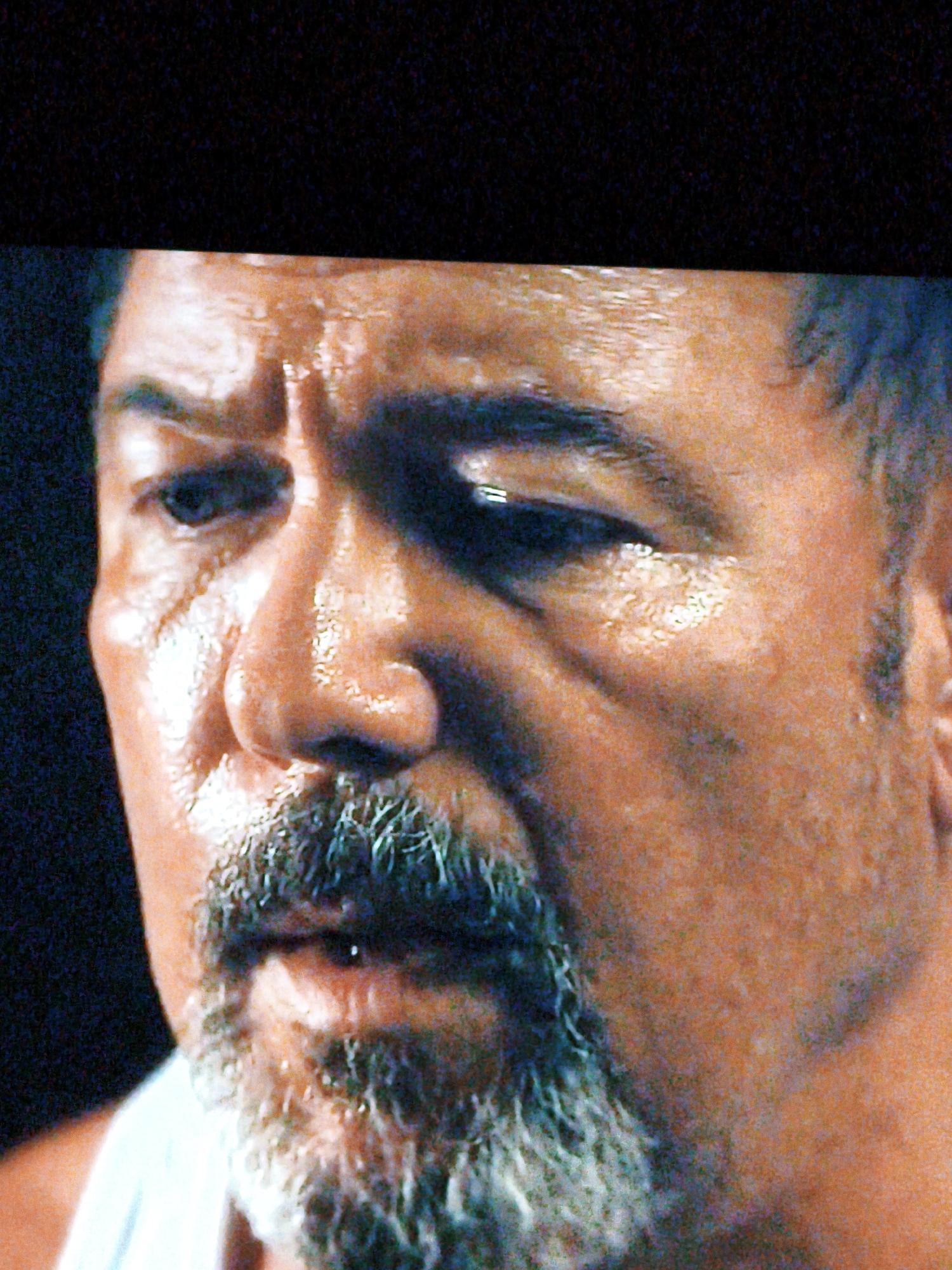 The image is a close-up, slightly blurry, and grainy photograph of a man's face, from the nose up. He has dark skin, a prominent nose, and a graying beard and mustache. His eyes are dark and appear to be looking slightly to the left of the camera. The lighting creates highlights on his forehead and cheeks, suggesting sweat or oil on his skin. The background is dark and indistinct. The image does not contain any visual information that can be used to determine its location in Vladivostok, Russia.