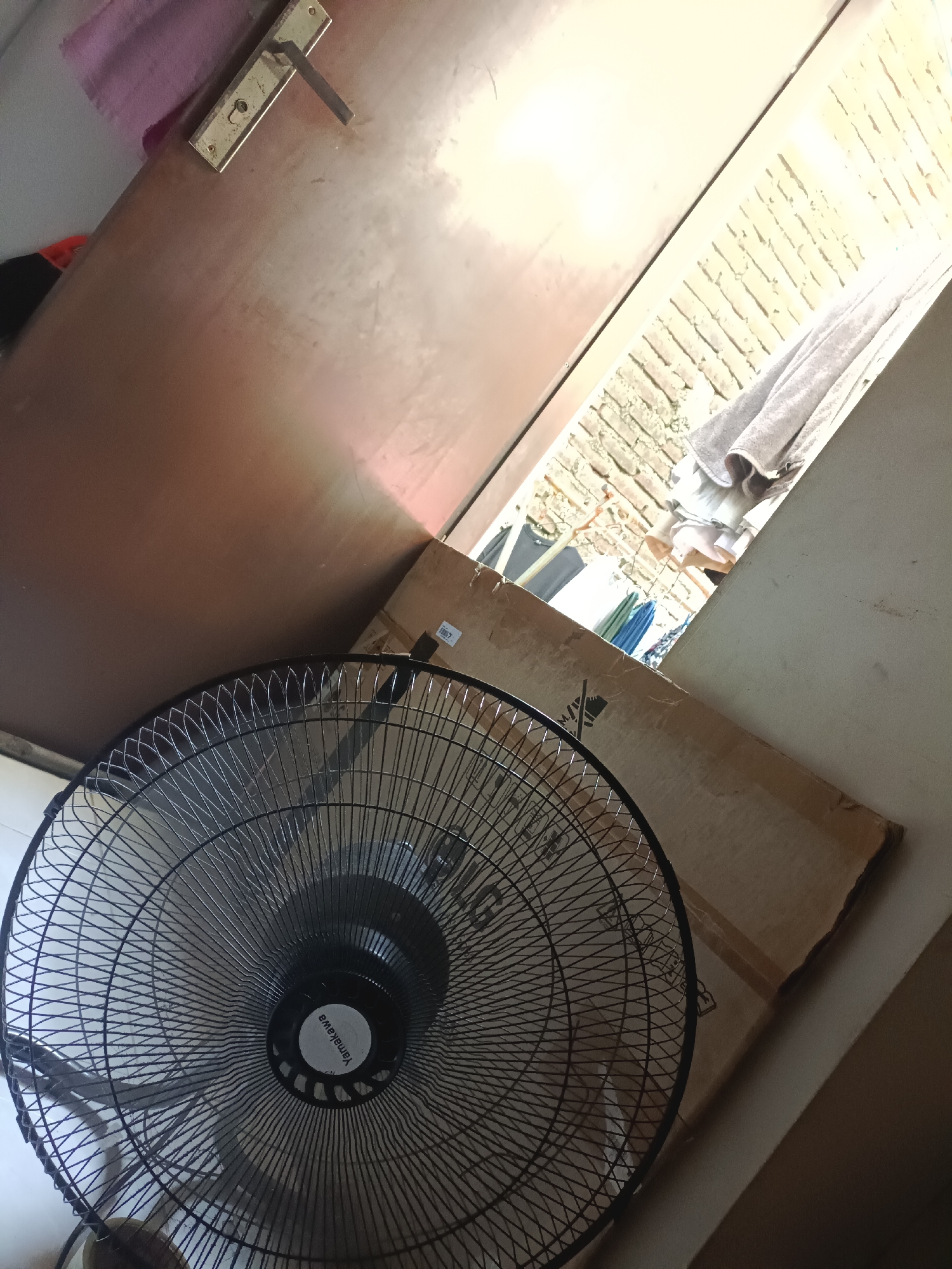 The image presents an indoor view looking outwards through an open doorway or large window. The scene is dominated by a large, black standing fan in the foreground, positioned towards the lower left. The fan features a metallic grille, and the brand name "Yamakawa" is clearly visible on its central hub.
To the left of the opening, a dark brown door or wooden panel is visible, partially open. A gold-colored door handle and lock assembly are affixed to this surface, with a small pink cloth draped near it.
On the right side of the doorway, a large, light brown cardboard box is leaning against an interior wall, partly obscuring the view of the exterior. Beyond the opening, a brightly illuminated outdoor area is visible, characterized by a light-colored brick wall. Several items of clothing, including a grey towel and other garments, are seen hanging on a line or rack, suggesting a laundry area or backyard.
The strong, natural light coming from outside indicates it is daytime, likely sunny. The presence of the fan suggests the interior might be warm. The setting appears to be a domestic space. No people are present, and no specific activity is actively taking place. The scene conveys a sense of everyday life.
