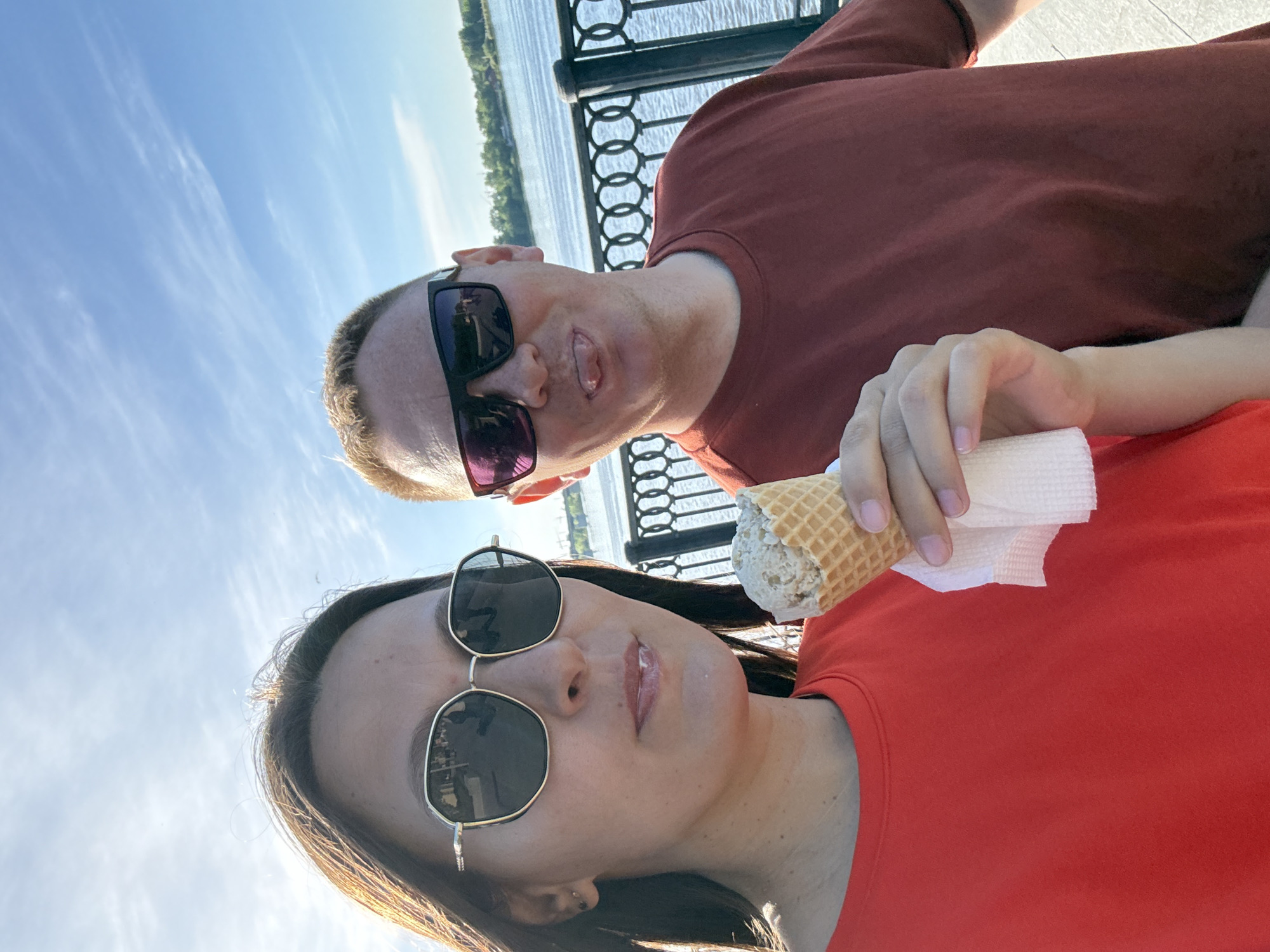 A selfie shows two people outdoors. The woman is in the foreground, wearing sunglasses and holding an ice cream cone wrapped in a napkin. She is wearing an orange t-shirt. Behind her is a man, also wearing sunglasses and a reddish-brown t-shirt. The background shows a metal railing, a body of water, and trees. Based on the architecture and landscape, the location is Perm, Russia.