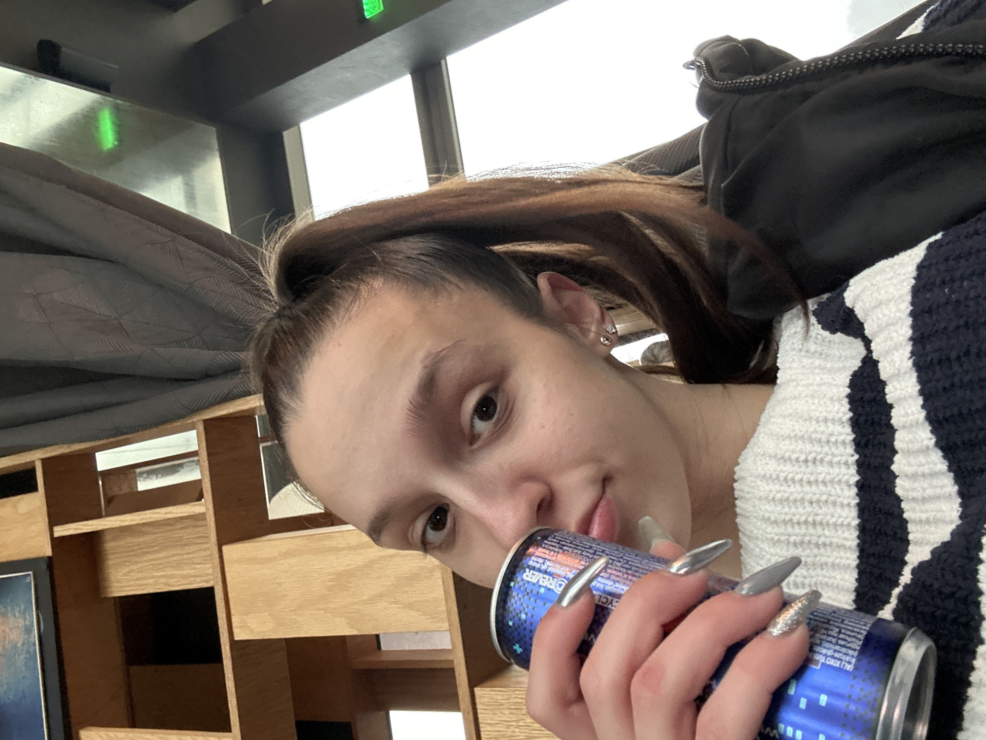 A young woman with brown hair in a high ponytail and long, metallic-colored artificial nails is holding a blue can, possibly an energy drink. She is wearing a white and black striped sweater and has a black jacket draped over her shoulder. The background features a wooden bookshelf and a window. The location is likely indoors and is stated to be in Varna, Bulgaria.
