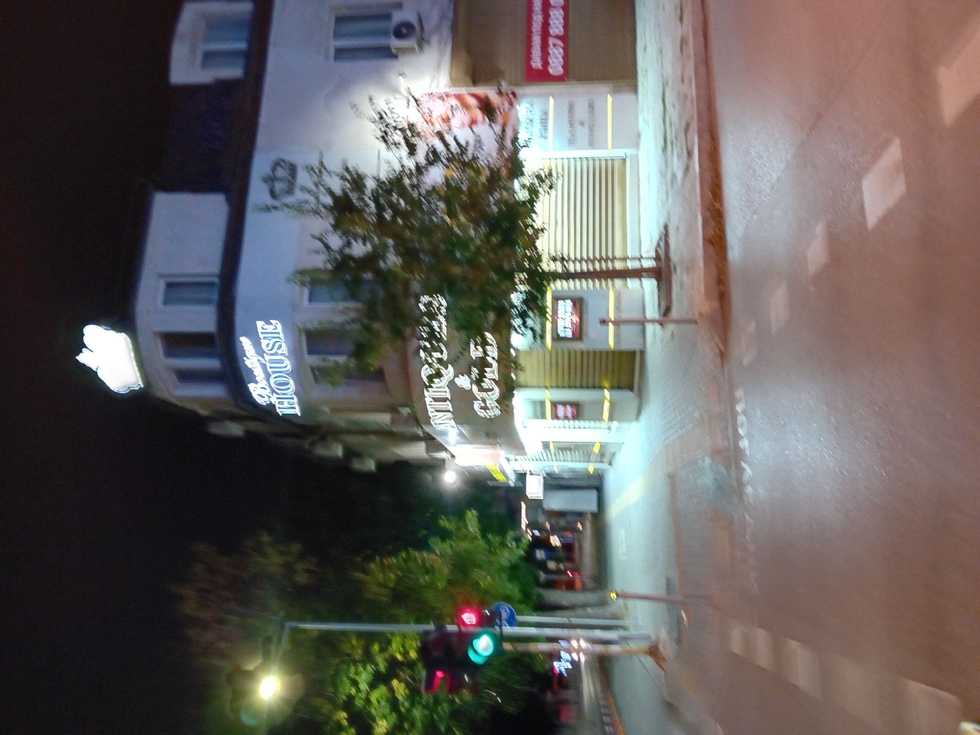 A blurry, nighttime image captures a street scene in Varna, Bulgaria. A traffic light shows a green signal. The facade of a white building is illuminated, featuring a neon sign that reads "Boutique HOUSE" with a crown emblem above it. Another sign displays "ANTIQUES & COFFEE." A small tree stands in front of the building. To the right, a red advertisement banner is visible, displaying a phone number: "0887 888 0." The street and sidewalk are wet, reflecting the ambient light.