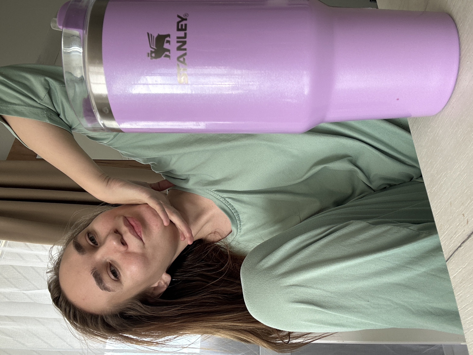 This image, taken in Kharnavy, Russia, features a woman and a large purple Stanley tumbler, rotated 90 degrees counter-clockwise. The woman, with long brown hair, is positioned on the lower left of the frame, looking towards the camera with a neutral expression. Her right hand is gently placed near her chin and neck. She is wearing a light green, possibly loungewear, top and matching pants, suggesting a relaxed indoor setting.
A prominent purple Stanley tumbler dominates the upper right portion of the image. The brand name "STANLEY" is clearly visible in gold lettering, along with its characteristic winged bear logo and a registered trademark symbol (®). The tumbler has a clear lid with a purple accent. A small, faint red dot is visible on the side of the tumbler.
The background includes natural light coming from a window covered by a light-colored curtain and a heavier brown curtain. A light-colored wooden surface, possibly a desk or table, is seen in the foreground and to the right. The overall impression is of a casual, domestic environment during daytime.