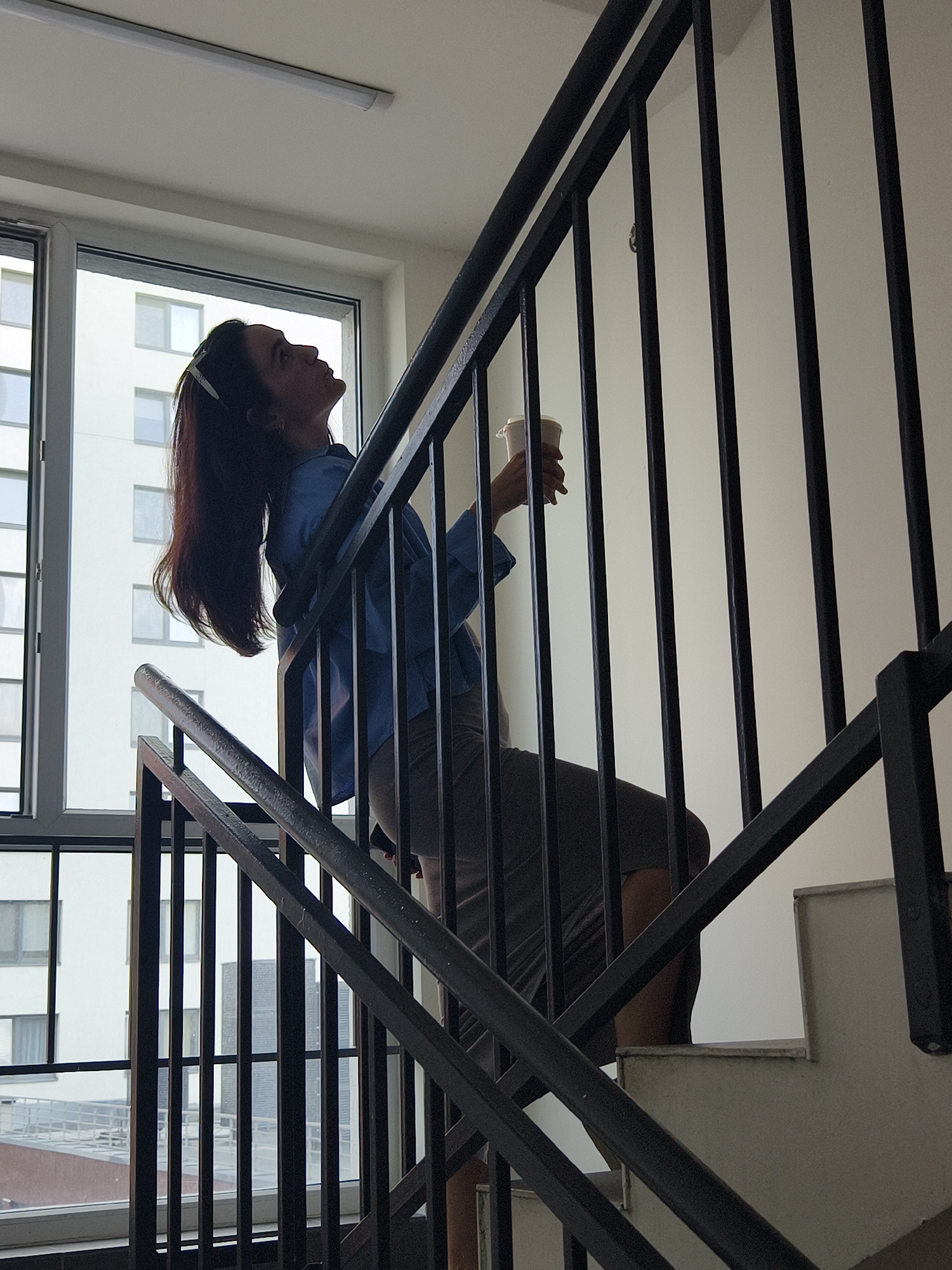 An image captured in the city of Tyumen, Russia, displays a female ascending an interior staircase. The subject, a woman with long, dark hair secured by a light-colored clip on her right side, wears a blue long-sleeved shirt and dark trousers. She is positioned on an upper step, looking upwards towards the ceiling, while holding a white disposable cup with a lid in her right hand. The staircase features black metal railings with vertical balusters and horizontal supports, and the steps are composed of light gray or off-white material. A large, multi-pane window with white frames dominates the left side of the scene, providing natural illumination. Through the window, several modern, white multi-story residential buildings with numerous windows are visible in the background, along with a darker, taller structure further in the distance. The interior walls and ceiling are plain white, with a single, linear fluorescent light fixture mounted centrally on the ceiling.