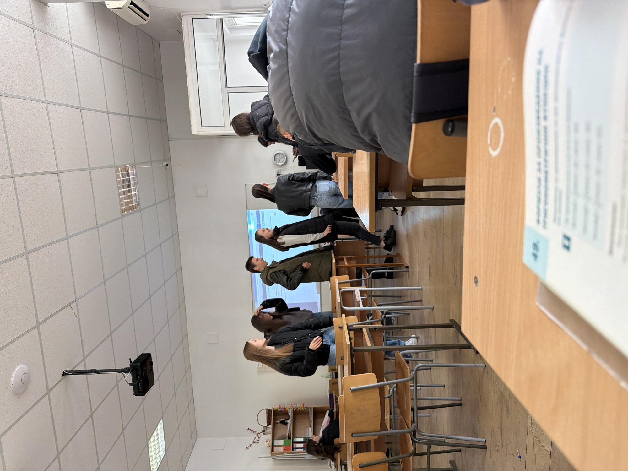 A classroom in Panagyurishte, Bulgaria, shows students standing and engaging in an activity facing a white screen. The room is furnished with desks and chairs, and shelves are situated near the back of the classroom. The lighting is provided by ceiling-mounted fixtures. There is an air conditioner is visible in the upper left corner of the frame. The photograph is taken at a tilted angle.