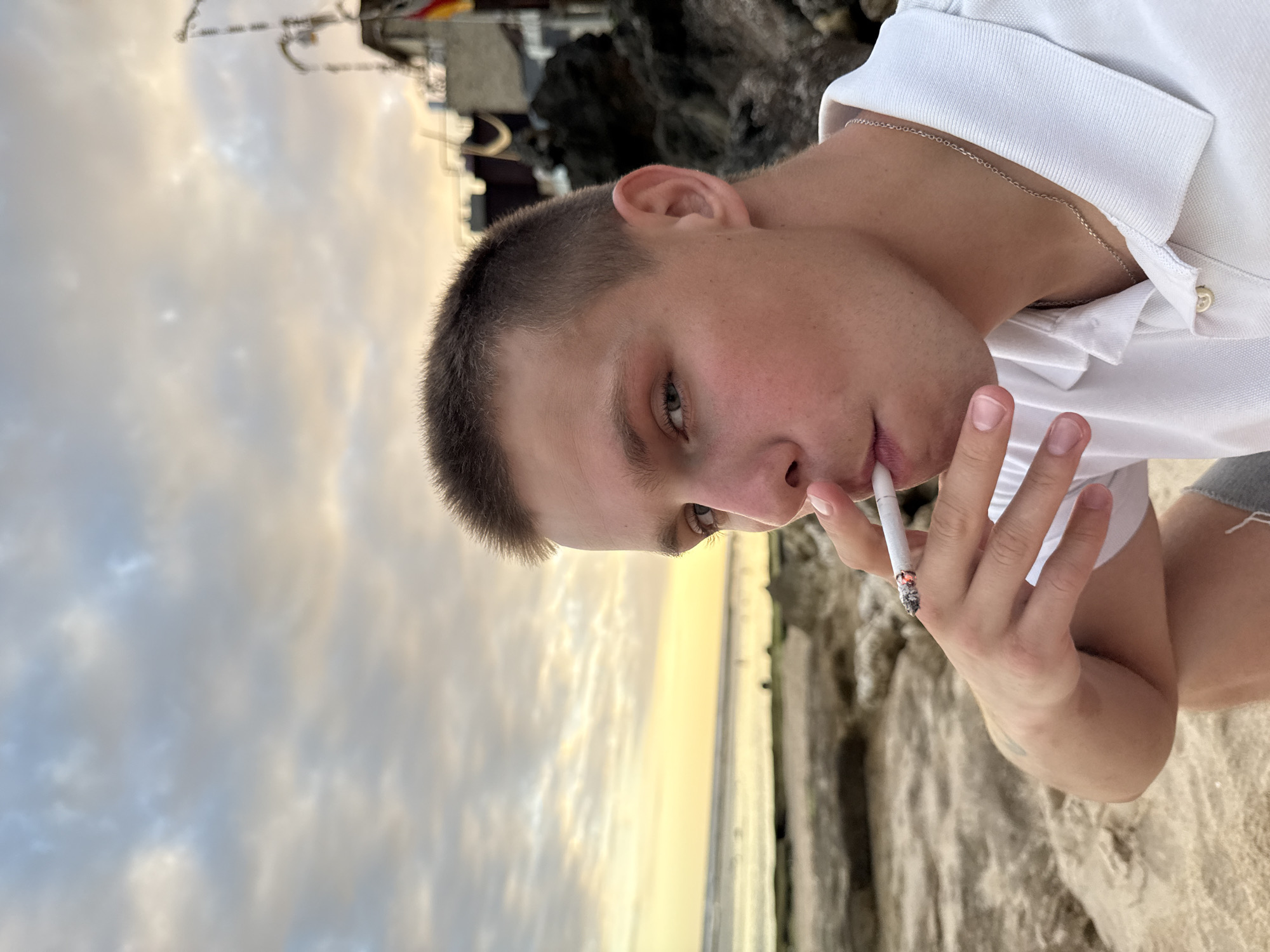 A fair-skinned young man with short, light-brown hair is smoking a cigarette. He is wearing a white polo shirt and a silver chain necklace. The background includes a beach and a building. The image was taken in Kuta Selatan, Indonesia.