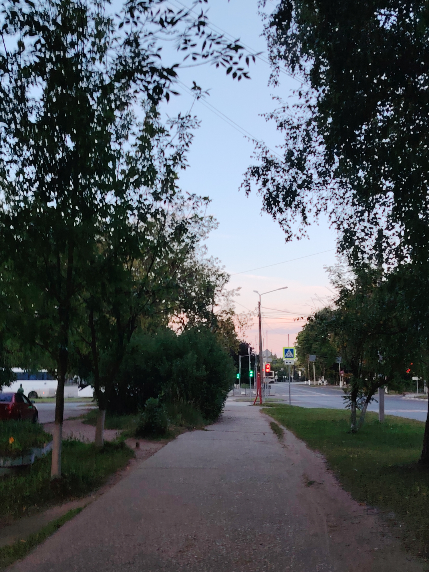 The image depicts a street scene in Konakovo, Russia, during twilight. A paved sidewalk runs through the foreground, bordered by grass and trees on either side. The sidewalk leads towards a street intersection with a pedestrian crossing marked by a blue and yellow sign. Traffic lights are visible, showing red and green signals. Several vehicles are present, including a white bus and a red car parked on the left side of the road. The sky is a soft blend of blue and pink, indicating the sunset or sunrise. The overall atmosphere is serene and quiet.