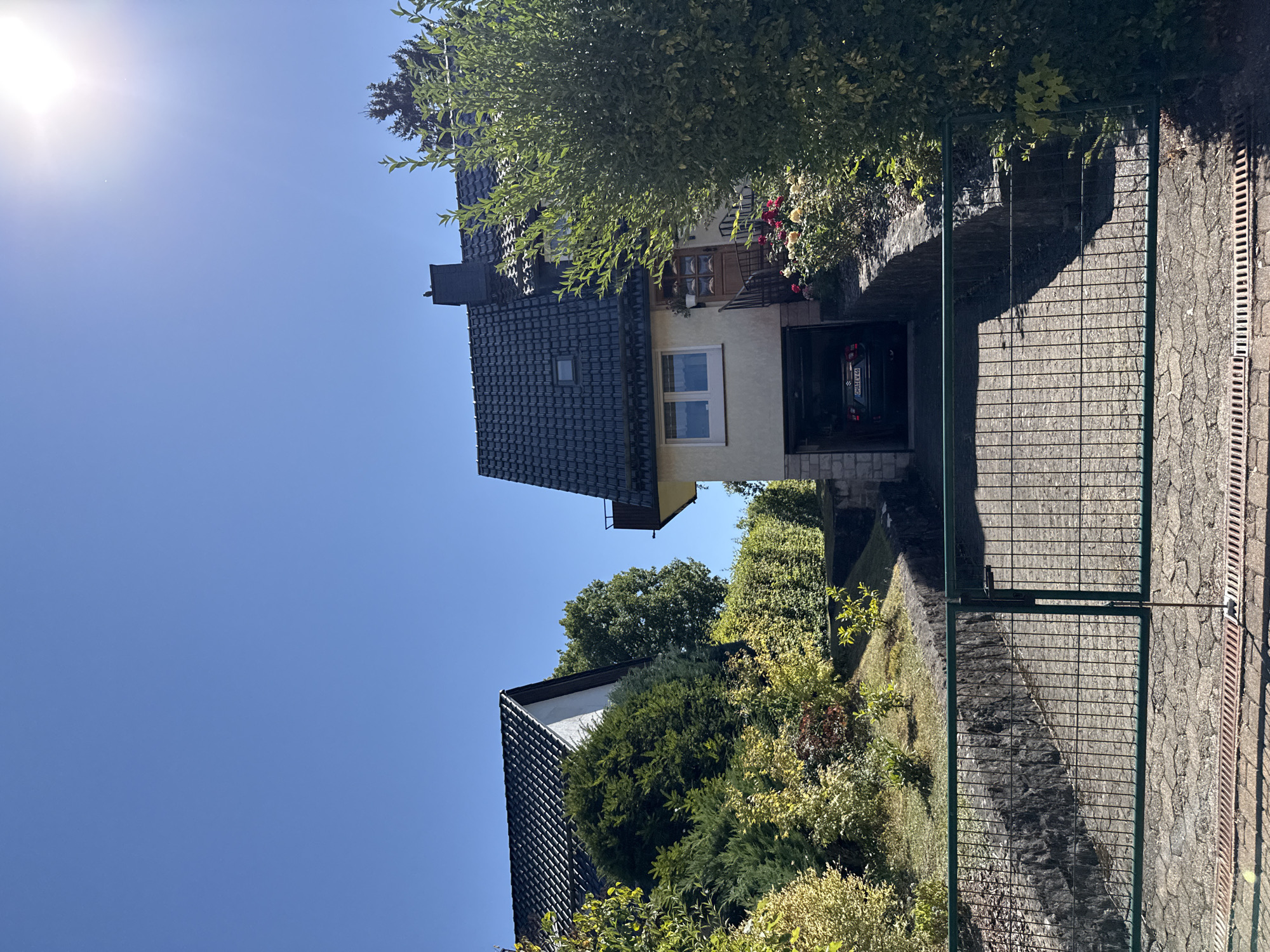 The image depicts a residential property in Bad Sachsa, Germany, on a clear, sunny day. The foreground features a green metal gate and a paved driveway. Beyond the gate, a sloped driveway leads up to a house with a dark-tiled roof, light-colored walls, and a garage entrance. A dark-colored car is visible inside the garage. Adjacent to the garage, a staircase leads to the main entrance of the house. The house is surrounded by lush greenery, including trees, bushes, and a flower bed with red and yellow roses. To the left of the main house, another house with a similar dark-tiled roof and a white facade is partially visible, nestled among dense green shrubs and trees. The sky is a clear, bright blue, with the sun prominently shining in the upper right corner, casting a bright glare. The overall scene is one of a well-maintained suburban home.
