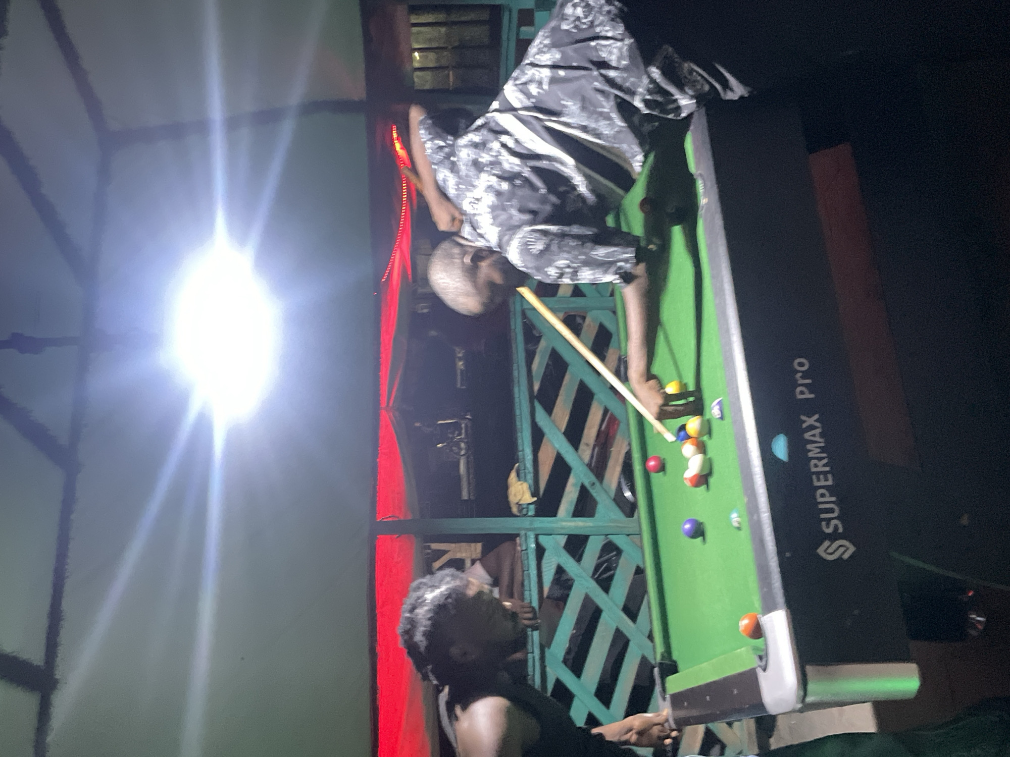 In a dimly lit outdoor setting, likely in Lokoja, Nigeria, two men engage in a game of pool. The older man is seen leaning over a "Supermax Pro" pool table, cue in hand, focused on the billiard balls. He's dressed in a patterned shirt, and the table is in play. The younger man, wearing a dark sleeveless shirt, watches the game. The setting seems informal with a partially visible wooden and metal structure behind the players, illuminated by a bright light source in the background, which could be a street lamp or security light. Red LED lights accent the structure. The scene captures a casual moment of leisure under the evening light.