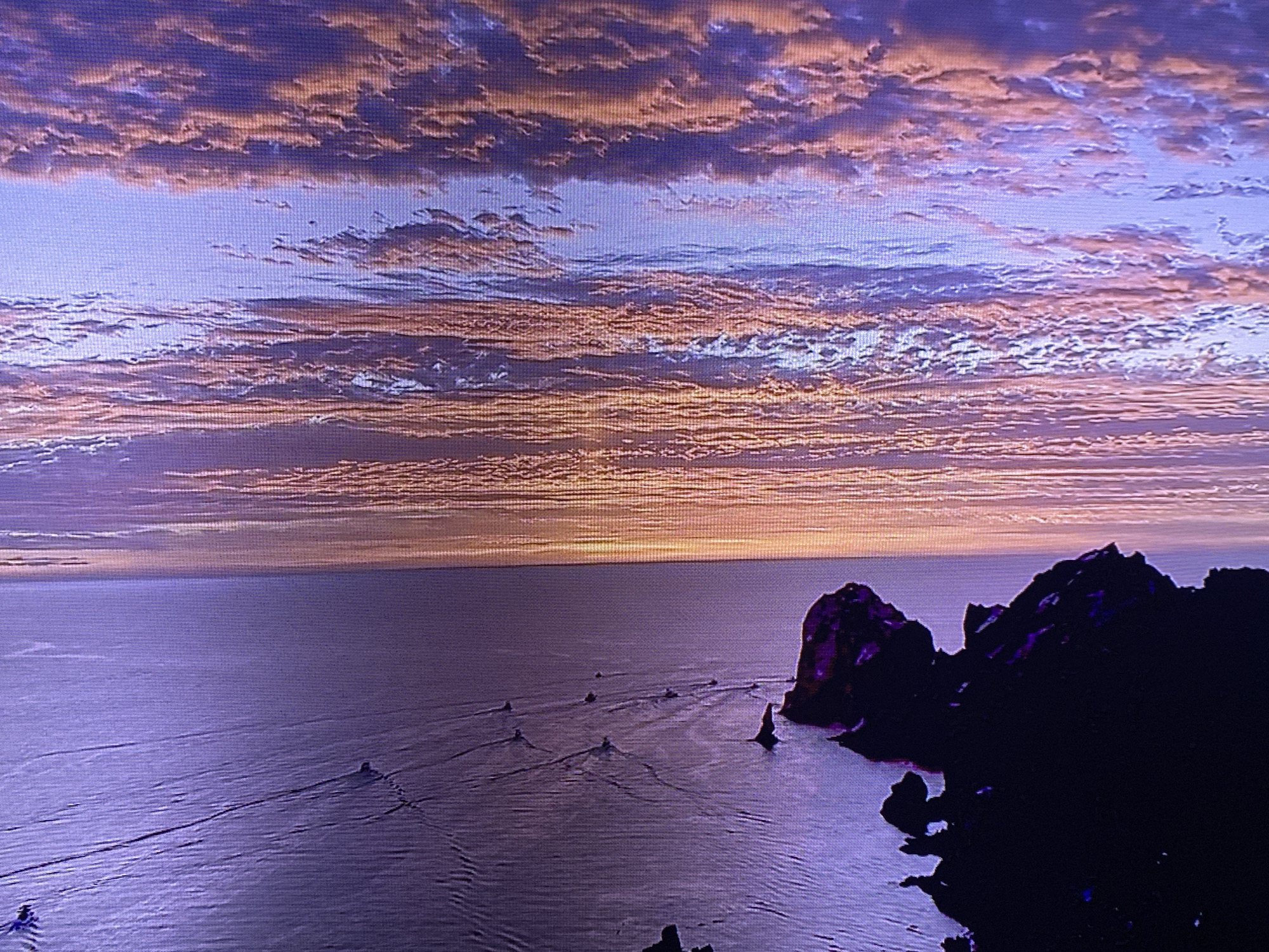 A wide shot shows a serene seascape at dusk. The sky is filled with streaks of pink, orange, and purple clouds, hinting at the last rays of the sun. Below the sky, a calm, purple ocean stretches out. Several small boats are visible, each leaving a distinct wake on the water, moving from left to right. In the foreground, dark, silhouetted rocky outcrops jut out from the water, framing the right side of the scene. The overall impression is one of tranquility and natural beauty.