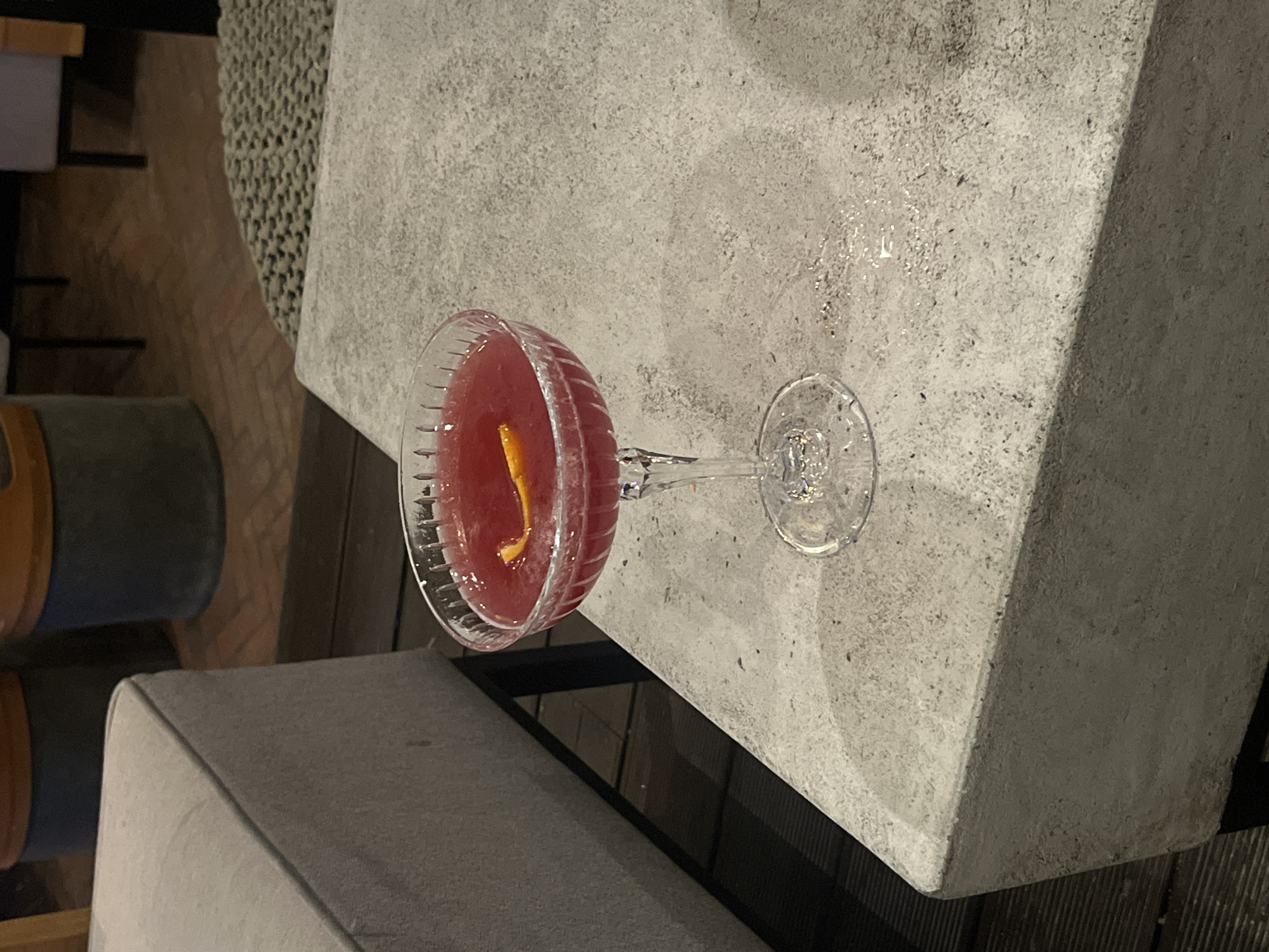 A close-up, overhead shot shows a bright red cocktail in a coupe glass with a fluted rim, garnished with a thin strip of orange peel. The glass sits on a textured concrete table, which is placed outdoors on a wooden deck. To the left of the table, a portion of a light gray upholstered bench is visible. In the background, blurred elements suggest a dimly lit outdoor seating area with large planters and chairs. The scene is devoid of people. The ambient lighting appears to be low, possibly evening.