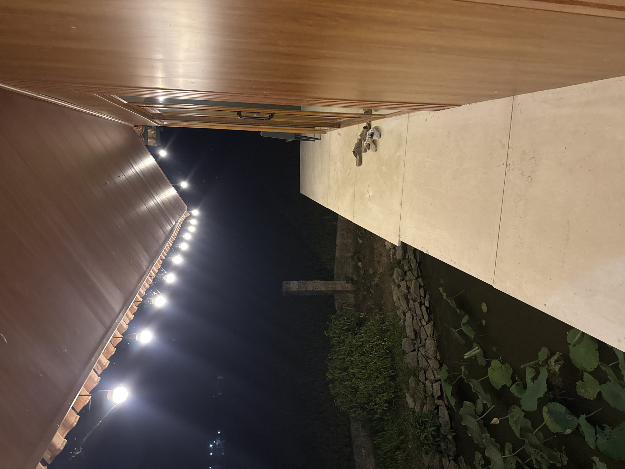 The image depicts an outdoor scene at night in Hà Giang 1 Ward, Vietnam. A lit building with a brown roof and string lights is visible on the left side of the frame. Adjacent to the building is a concrete walkway with a pair of shoes and sandals. To the right of the walkway is a rock wall, vegetation, and a body of water with lily pads. The background is dark, with faint lights visible in the distance.