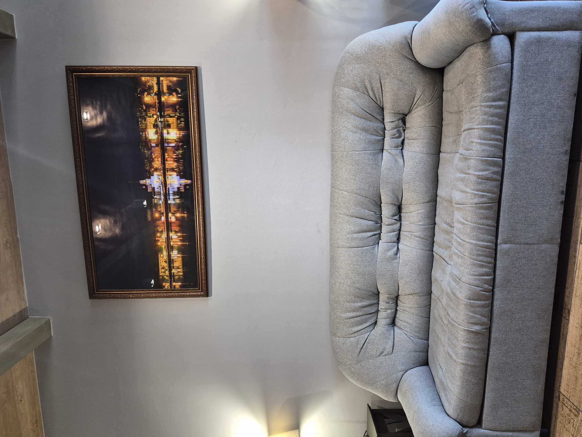 A framed photograph hangs on a gray wall beside a gray sofa. The photograph depicts a nighttime cityscape with illuminated buildings and reflections in the water. The sofa is made of a soft, tufted material and has a rounded shape. A wood-paneled wall is partially visible on the left side of the photograph. No people are visible. The city of Votkinsk, Russia, cannot be confirmed based on the visual information provided.