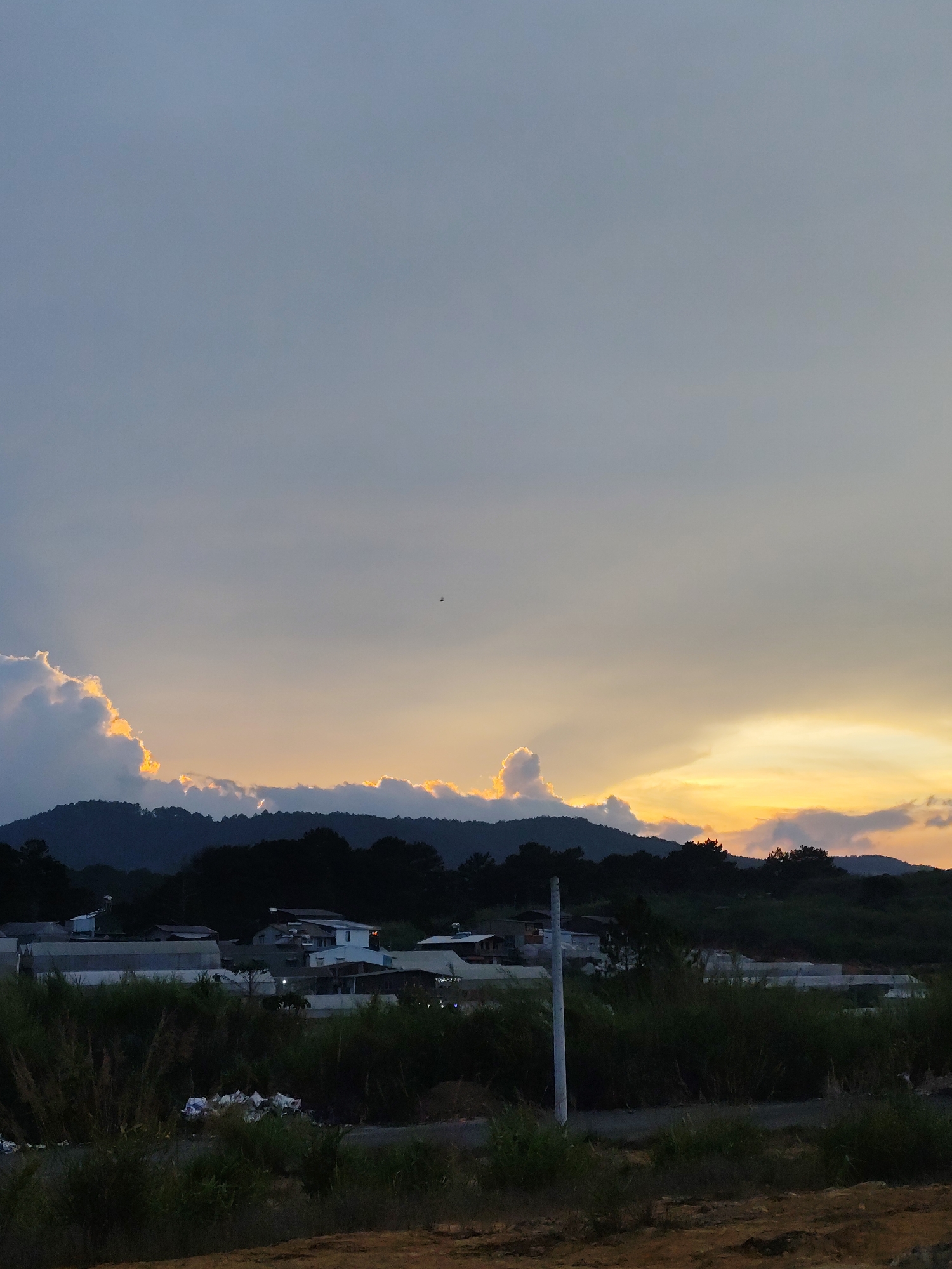 The image shows a landscape scene in Da Lat, Vietnam, at dusk. The foreground is a dirt field with vegetation. In the midground are buildings, possibly residential and agricultural structures, and a power pole. A line of dark green hills forms the horizon. Above the hills, the sky is filled with clouds, with colors ranging from gray to golden orange. A small bird is visible in the sky.