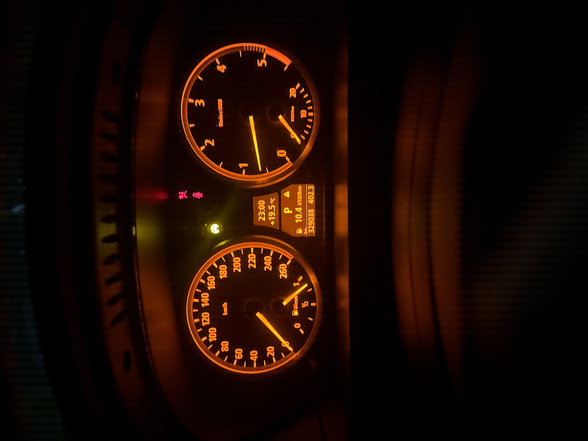 The image shows an illuminated dashboard of a vehicle. The speedometer indicates approximately 30 km/h. The tachometer reads below 1000 RPM. The temperature is 19.5 degrees Celsius. The dashboard display shows 23:00. The distance is 329038 and the distance to the next destination is 402.3. The car is in park and consumes 10.4 l/100km. It also shows the fuel tank is half full and the car is running on diesel. There are also 3 lights on. There is not enough information to determine the location.