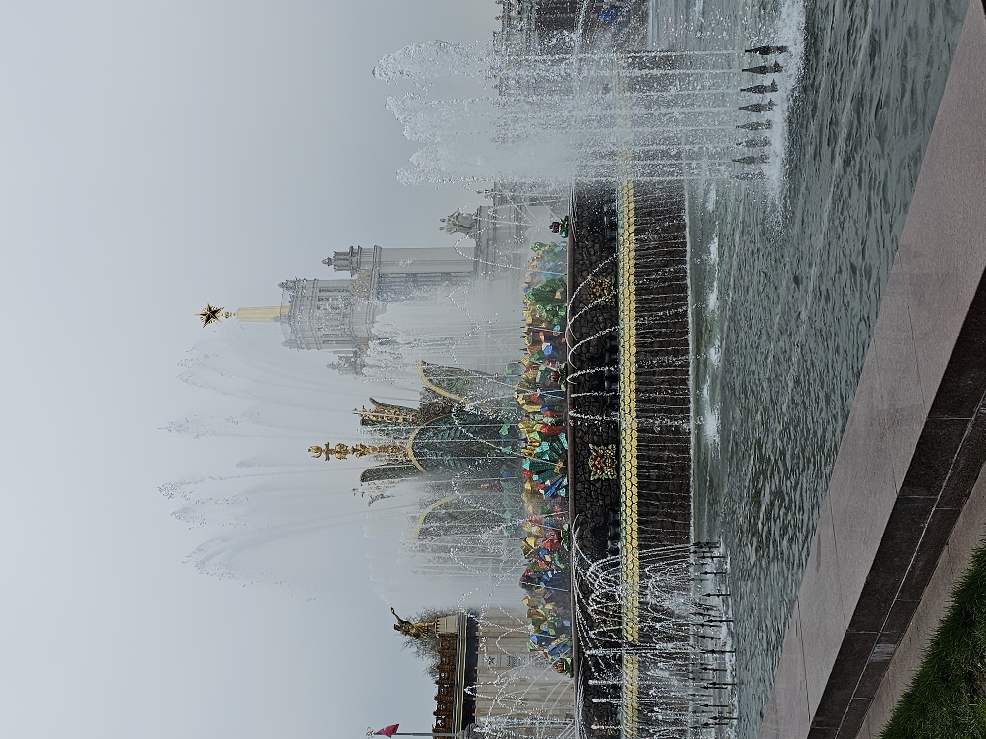 This image captures a wide shot of a majestic, active fountain and a grand architectural landmark under an overcast sky in Moscow, Russia. The scene is dominated by a large, ornate fountain, likely the Friendship of Peoples Fountain at VDNKh. Multiple powerful jets of water spray upwards and outwards, creating dynamic patterns and a misty effect. The fountain's central structure is richly adorned with golden embellishments and a vibrant array of colorful, faceted elements, possibly mosaics or artificial jewels, arranged in geometric patterns. The water in the fountain basin appears turbulent from the spray.
In the background, a palatial, light-colored building with intricate architectural details stands, partially obscured by the fountain's spray and atmospheric haze. This prominent structure features a towering golden spire, which is crowned by a distinctive five-pointed star. Other buildings are faintly visible further in the distance, contributing to the grand scale of the setting.
The foreground shows the edge of the fountain's dark-colored basin, with a small patch of green grass visible on the far right. The sky is uniformly grey and overcast, indicating a cloudy day, and the lighting is diffused. No people are visible in the frame, and there is no visible text. The overall atmosphere is one of stately beauty and historical grandeur.