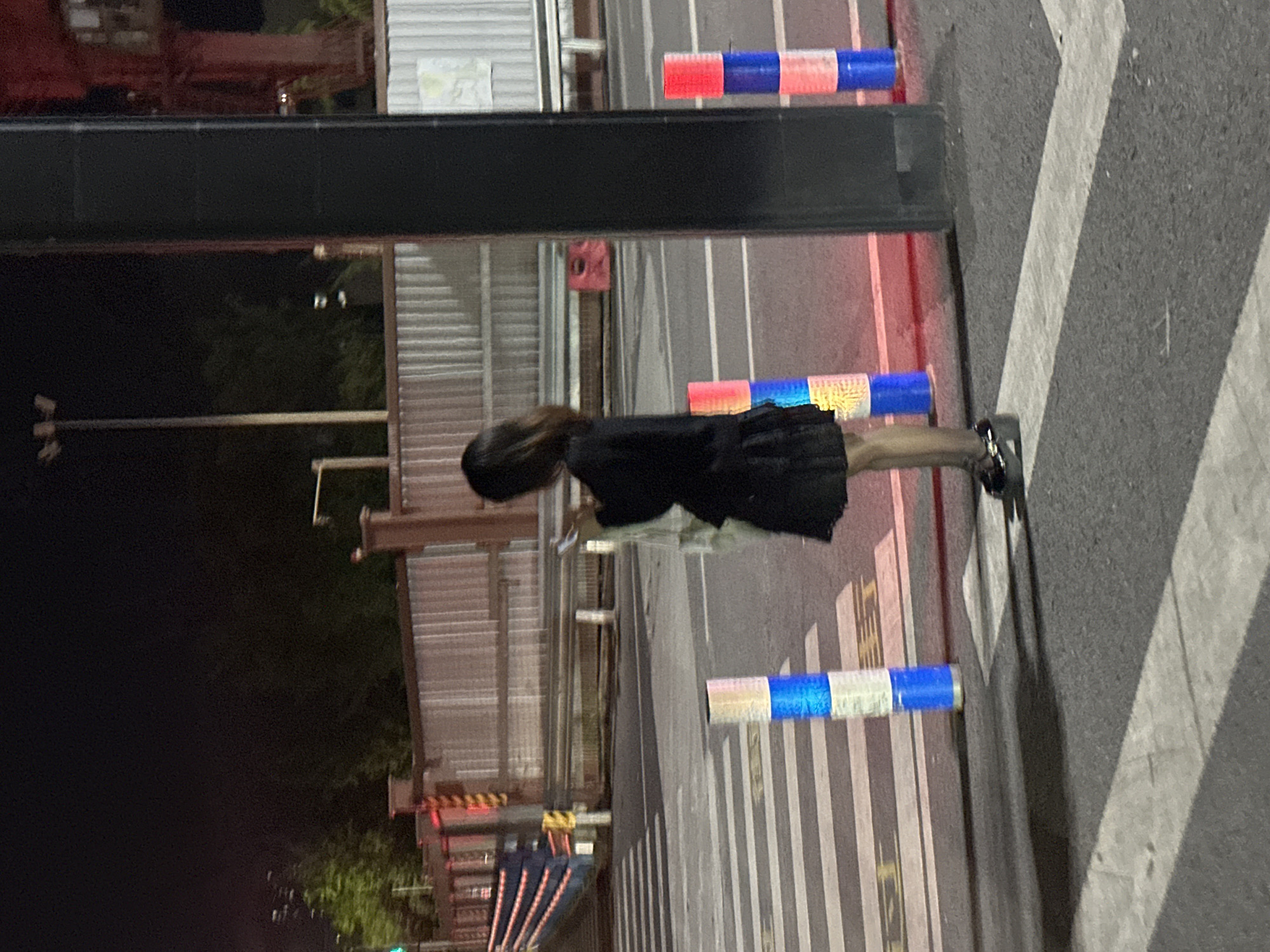 A person stands on a zebra crossing at night, looking at a mobile phone. The person is wearing a black dress and platform shoes. There are traffic bollards with blue and red lights on the crossing. In the background, there is a fence and some construction equipment. The scene appears to be in a city, possibly in the Chongqing High-tech Zone, China, given the characteristic red and yellow striped traffic barriers and the overall urban lighting. The lighting is artificial, casting shadows on the road.