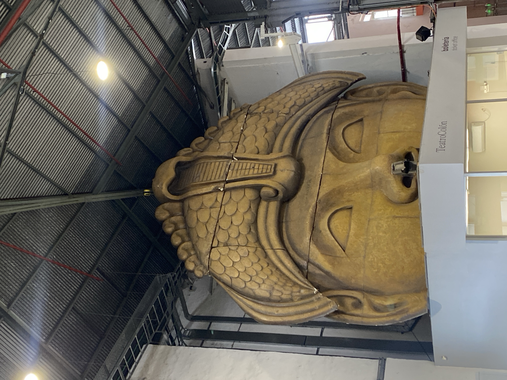 The image displays a massive, golden-colored, intricately carved head, appearing to be a large theatrical prop or statue, situated within an indoor industrial environment. The head is adorned with a detailed headdress featuring overlapping scale-like or feather-like textures, and it displays closed or downcast eyes with a serene expression. Its surface is a weathered metallic gold, showing visible seams and cracks, suggesting it is constructed from multiple pieces.
The setting features a high, open ceiling composed of corrugated metal panels, exposed metal trusses, and beams, with red pipes running overhead. Bright artificial lights illuminate the space. To the right of the head, a modern, light-colored structure with large windows is visible. A prominent vertical sign on this structure explicitly states "Teatro Colón" and "boletería / ticket office." This text confirms the location is associated with the famous Teatro Colón in Buenos Aires, Argentina, likely a backstage area, workshop, or storage facility for large props. No human activity is depicted; the scene presents a static display of this grand theatrical element.