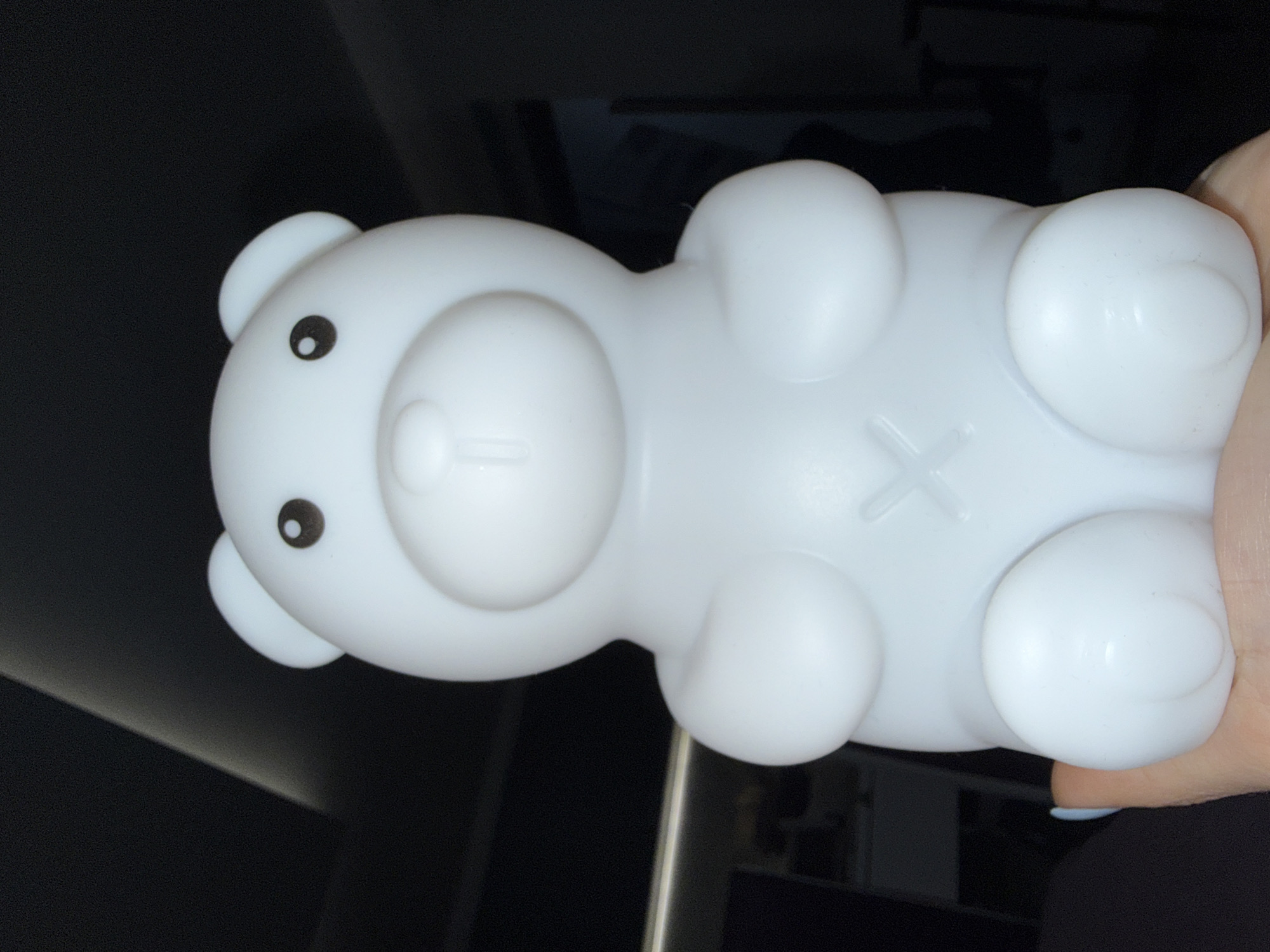 A white, plastic bear-shaped nightlight is held in someone's hand. The nightlight has black eyes, a circular nose, and an "X" shaped mark on its stomach. The background is blurry and dark. There is no evidence in the picture to suggest the image was taken in Plovdiv, Bulgaria.