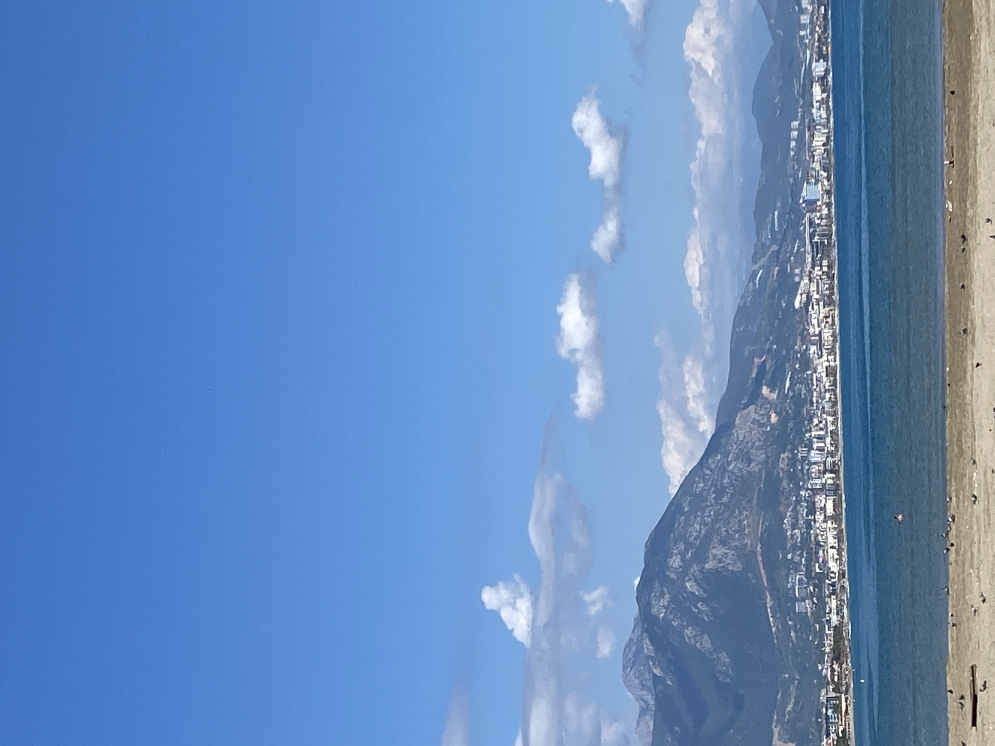 The image depicts a coastal scene in Alanya, Turkey. A clear blue sky dominates the left portion of the frame, punctuated by scattered, fluffy white clouds. Adjacent to the sky is a view of the mountains, which rise steeply, covered with vegetation and dotted with buildings indicating human habitation. At the foot of the mountain lies the city, with a dense concentration of buildings extending along the coastline. The turquoise waters of the Mediterranean Sea stretch out to the right of the city, and a sandy beach, populated with a few indistinct human figures, occupies the extreme right edge of the scene. The image appears to be taken on a sunny day, with high visibility allowing for sharp detail of the landscape.
