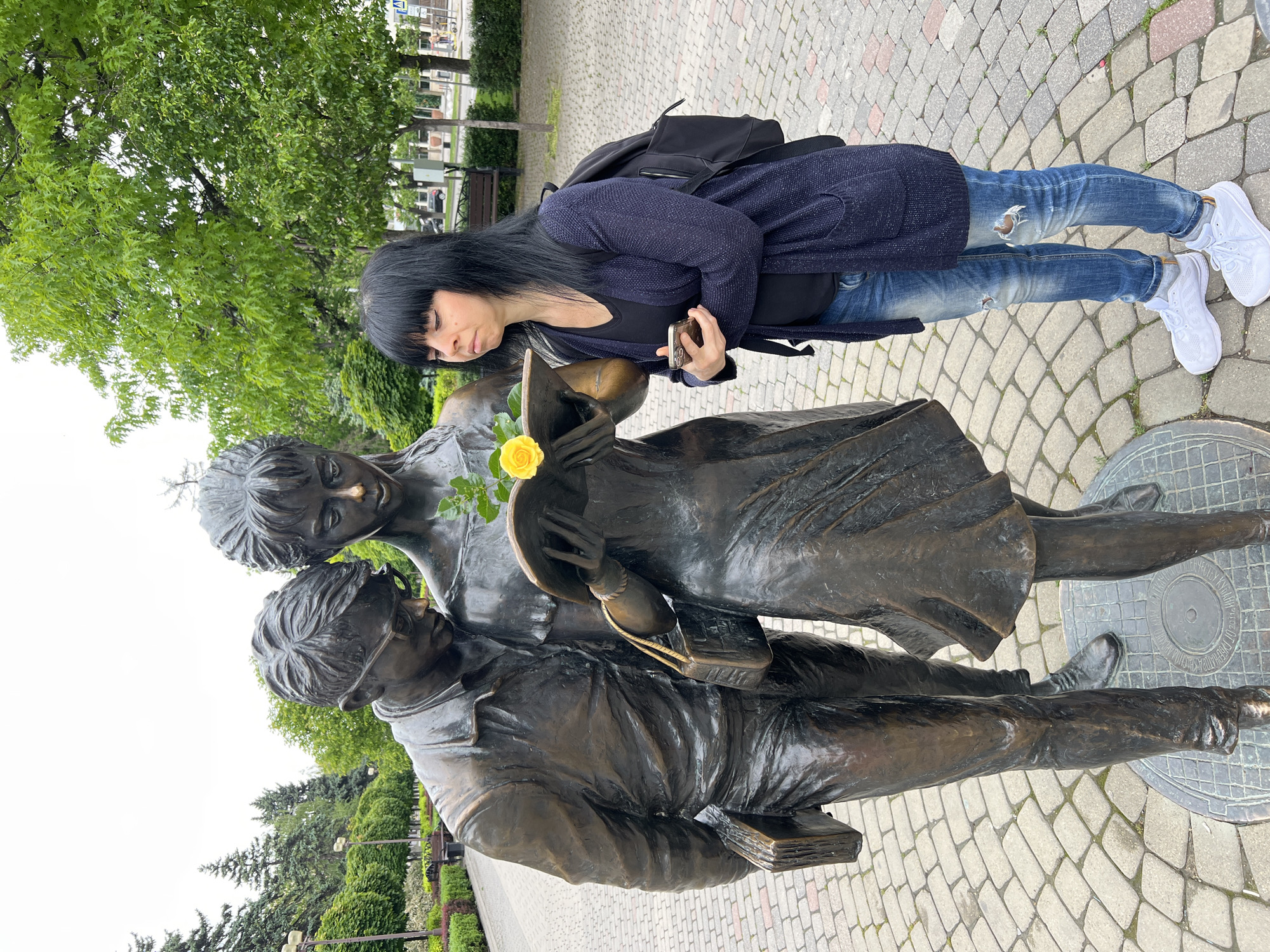 A woman in jeans and a dark cardigan poses beside a bronze statue in Krasnodar, Russia. The statue depicts two young people in Soviet-era attire. The girl is holding a yellow flower. The woman is holding a phone and looking down at the statue. The ground is paved with cobblestones. Trees and shrubs are visible in the background.