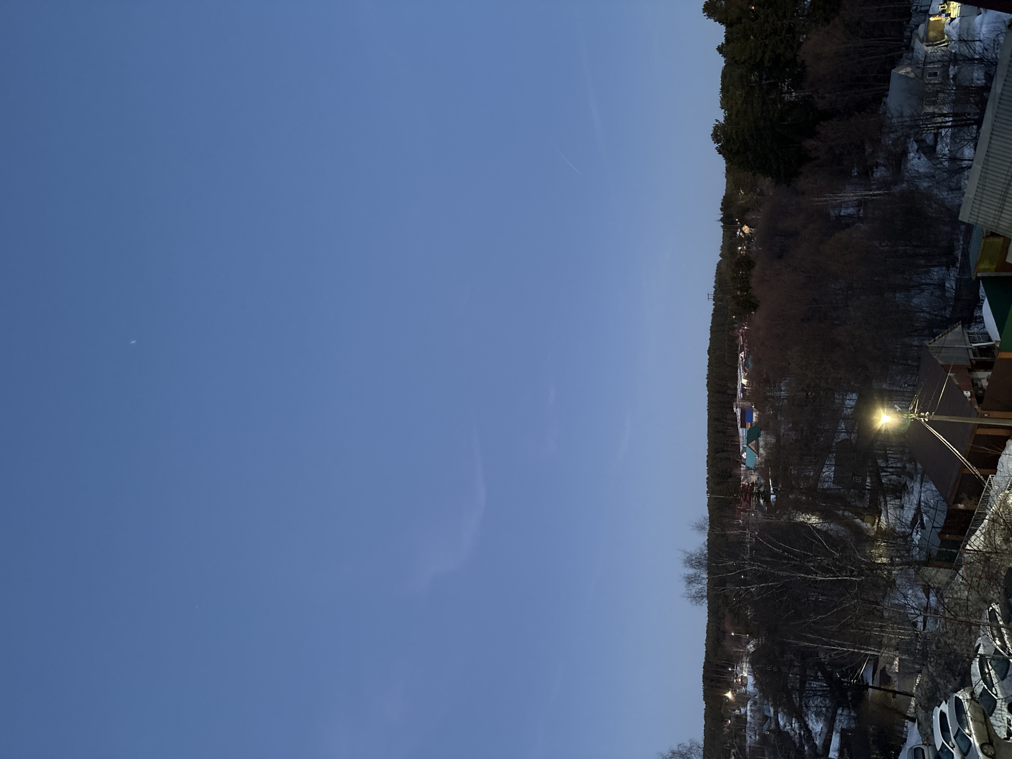 The image presents a view of the city of Chaikovsky, Russia, in what appears to be late evening or early morning. The sky is a clear, deep blue, dominating a large portion of the frame. A line of trees and buildings is visible along the horizon. A section of a building, a streetlight, and parked vehicles occupy the lower right corner of the frame. Snow is present on the ground amidst the trees.