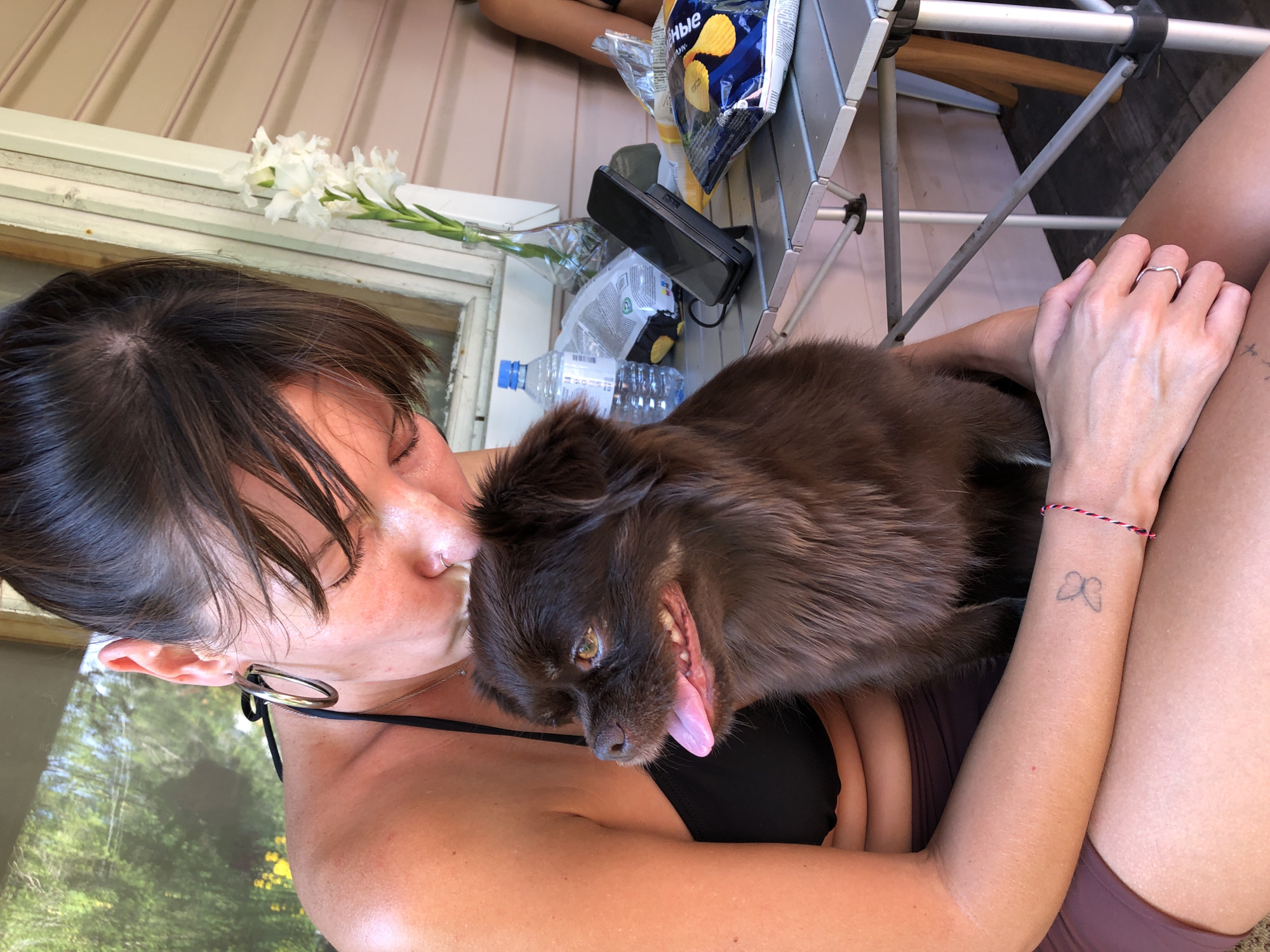 A woman with dark hair, wearing a black bikini top and maroon shorts, is holding a small, fluffy brown dog in her lap. She is leaning down to kiss the dog, who is looking to the left with its tongue out. The woman has large hoop earrings and a nose ring. On her left wrist, there is a small butterfly tattoo and a red and white string bracelet. She is also wearing a silver ring on her left index finger.
They are sitting on a porch or deck, with a table in front of them. On the table are a bottle of water, a vase with white flowers, a mobile phone on a stand, and bags of potato chips. The background shows greenery and a white wooden structure. The scene suggests a casual outdoor setting. The location of Красная Пресня Снт, Russia, is mentioned, but there are no specific visual cues in the image to confirm this precise location.