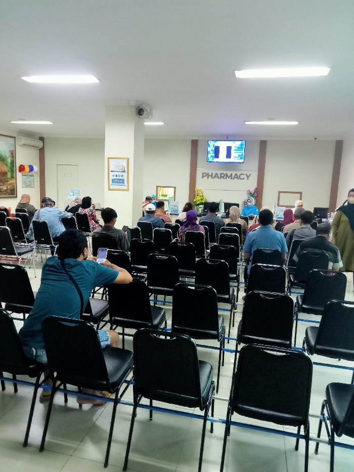 The image shows a waiting area in a building located at latitude -6.76, longitude 111.015 in Kecamatan Margorejo, Indonesia. The waiting area is filled with black plastic chairs arranged in rows. There are several people sitting in the chairs. Some of the people are looking at their phones while others are talking or looking around. A sign above the waiting area reads "Pharmacy". The walls are white and there are fluorescent lights above the waiting area. The floor is tiled and clean. There is a large window behind the waiting area, but it is not clear what is outside. There is a security camera in the ceiling. A person wearing a blue shirt is sitting in the chair and holding a cellphone. There is a woman wearing a blue hijab in the background, standing with another person in the distance.