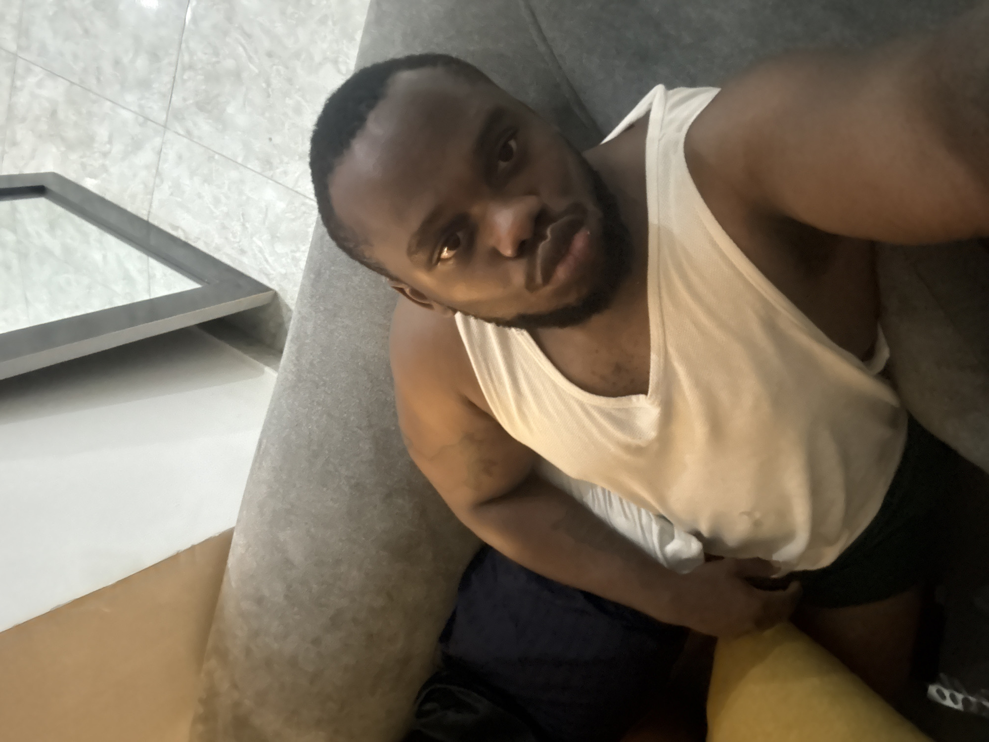 A person with dark skin and short, dark hair is sitting on a grey couch wearing a white tank top and dark shorts. The person is looking at the camera with a slight frown and pursed lips. The background includes a light-colored tiled floor with a partially visible framed mirror and some of the room's furnishings. No information relating to Iddo Tudunwada, Nigeria, is discernable.