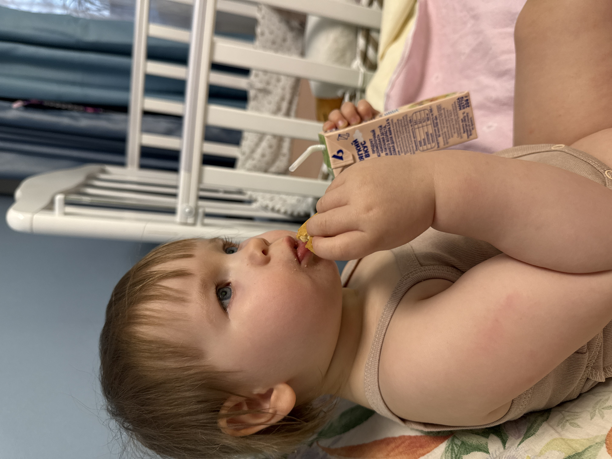 A toddler with light brown hair and blue eyes is lying on their side, eating a snack and holding a juice box with a straw. The text on the juice box is in Russian. They are wearing a light brown tank top. A white crib is visible in the background. The location appears to be indoors.