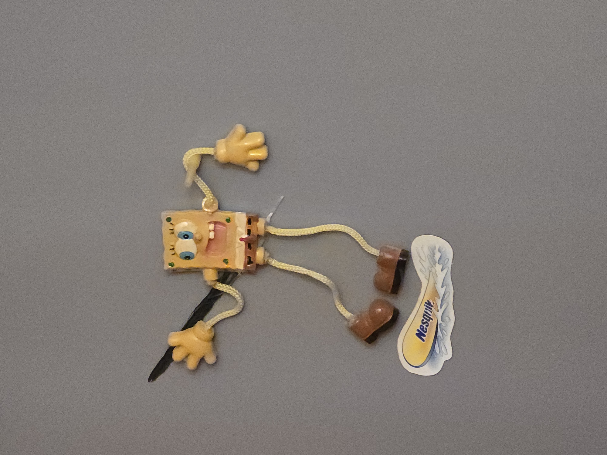 A toy figure resembling Spongebob Squarepants, made of a light brown material, is positioned on a gray surface. The figure has large blue eyes and a visible pink mouth. It is connected to yellow, rope-like limbs that are attached to light brown, boot-like feet. A black cord extends from the figure's lower body downwards. A white sticker with blue and yellow coloring, featuring wavy lines and the text "Nesquik" in cursive, is located to the right of the toy. The lighting suggests an indoor setting, with no indication of weather or time of day.