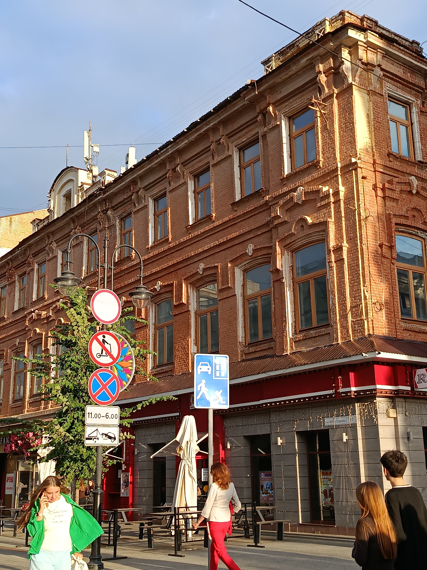 The image shows a street scene in Saratov, Russia, with a focus on a brick building and traffic signs. The building appears to be old, with ornate architectural details and multiple stories. Several people are visible walking on the sidewalk. There are traffic signs indicating parking restrictions and prohibitions on scooters. A tree is also present, and there are outdoor seating areas, suggesting a commercial or public space. The sky is visible and appears to be a clear blue, indicating daytime.