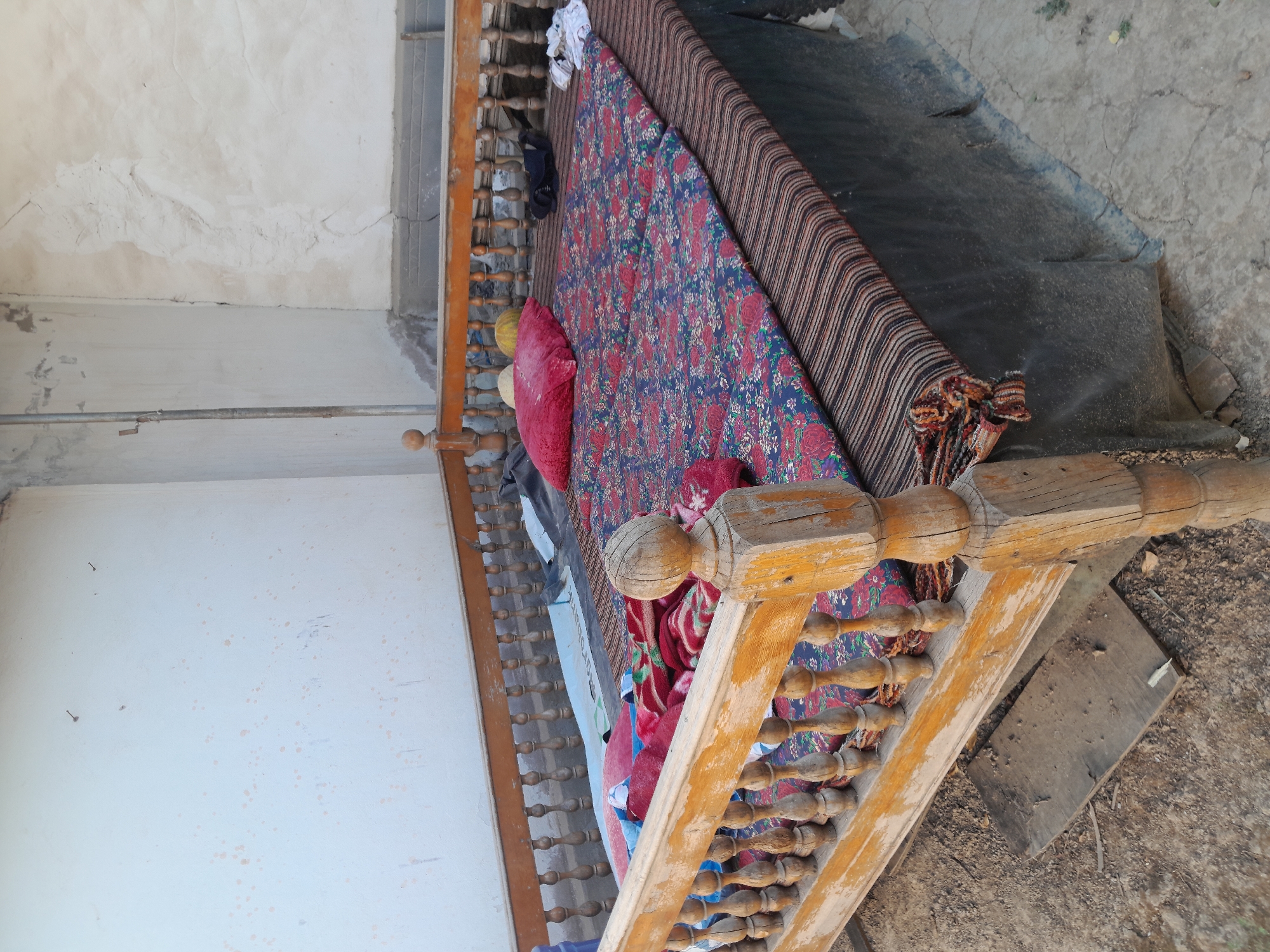 In the city of Unknown, Unknown, a wooden bed frame with turned spindles and weathered, light-brown wood is positioned outdoors. The bed is covered with a striped mattress and two floral-patterned mats. A red pillow sits on the mats, along with a red and white blanket. A pair of black pants and white fabric are draped over the side of the bed. The bed is on a dirt and gravel ground, with a cracked concrete patio visible. The background consists of a white, textured wall and a partial view of another wall with cracks.
