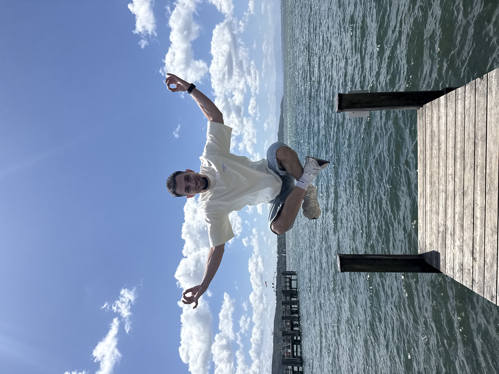 A young man with short dark hair and a beard is captured mid-air in a meditative pose, appearing to levitate over a wooden pier on a sunny day. He is wearing a light-colored t-shirt, denim shorts, and white sneakers. His arms are extended outwards, with his hands forming a gesture, possibly a mudra. The background features a calm body of water, a pier with wooden posts, and a distant shoreline with trees and mountains under a bright blue sky with scattered white clouds. The image is taken from a low angle, emphasizing the man's levitation against the expansive sky and water. The location is Starnberger See, Germany.