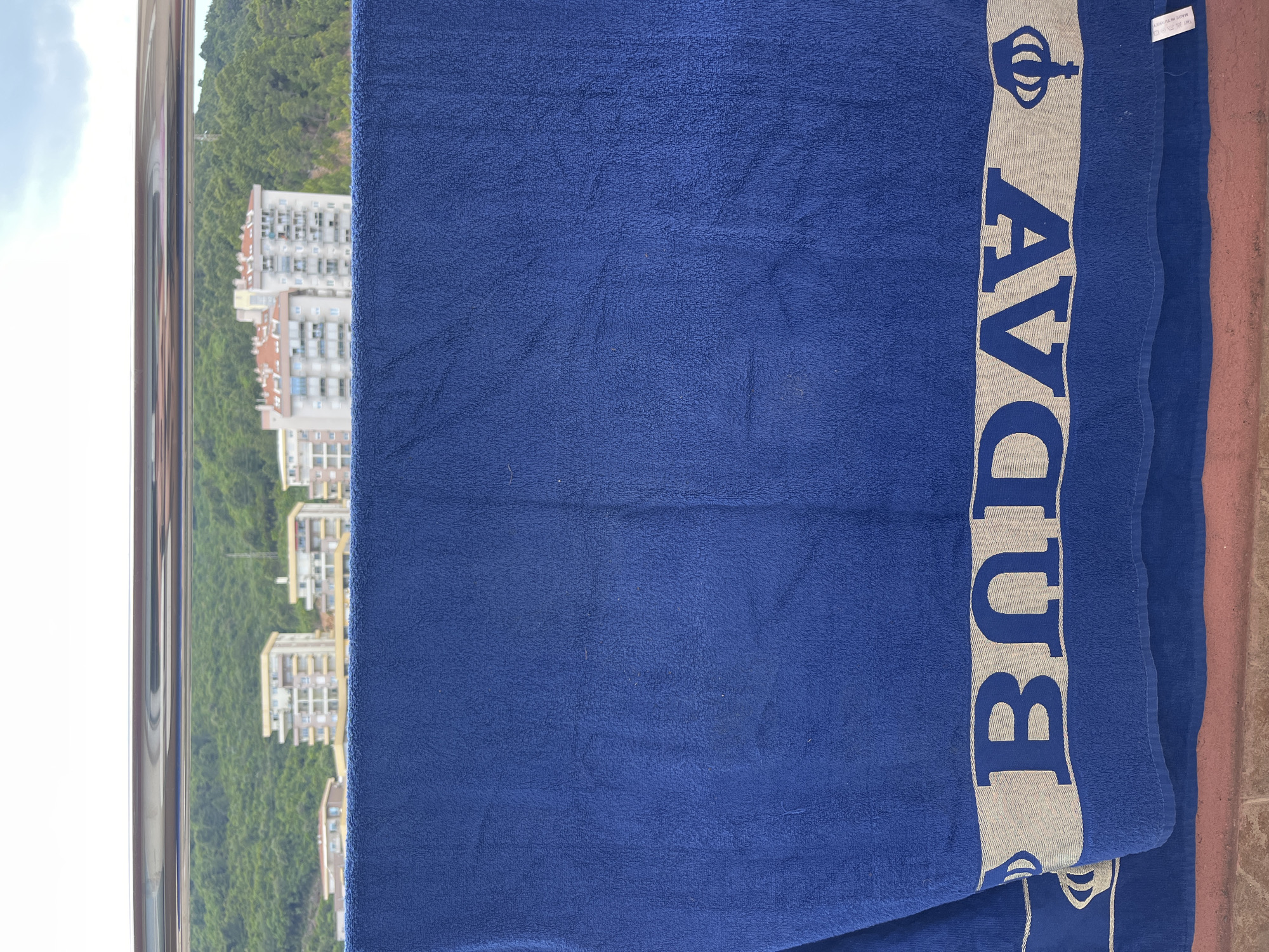 A blue beach towel is displayed vertically, predominantly covering the view. The towel has a vertical band on the right with the word "BUDVA" printed in large white block letters, accompanied by a crown symbol above the word. The towel is draped over a balcony railing. Outside the railing, the background reveals a cityscape with multi-story buildings of beige and white hues, nestled against a backdrop of a lush green hillside. The sky above is partly visible with white clouds. The city is Budva, Montenegro, identifiable from the text on the towel.