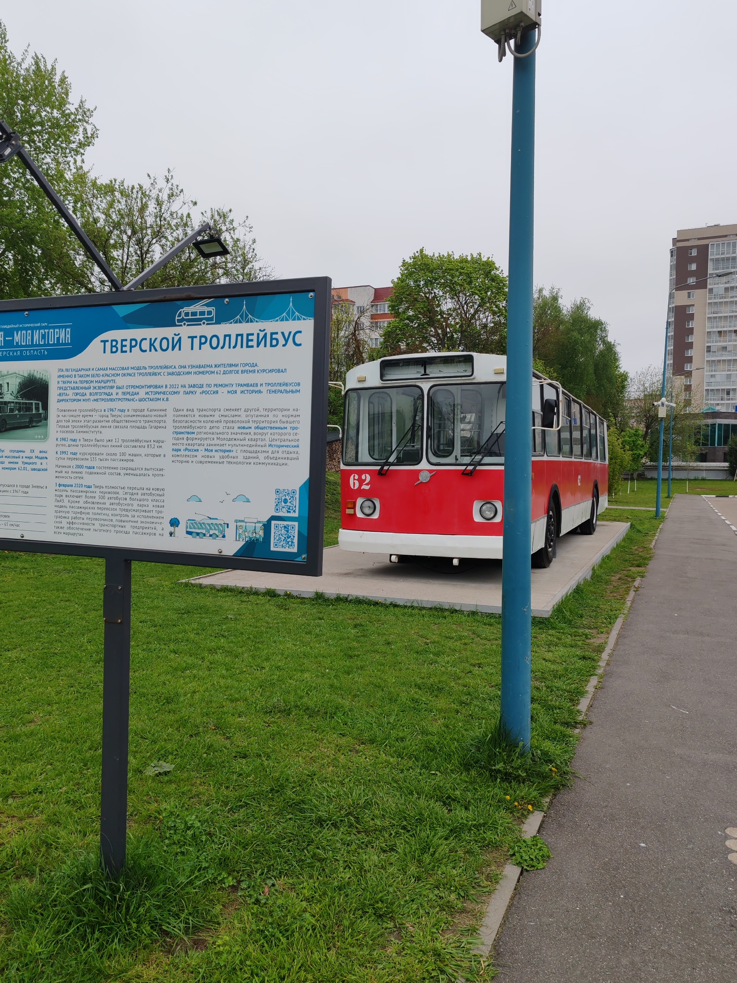 The image captures a static exhibit of a red and white vintage trolleybus, number 62, stationed on a concrete platform adjacent to a paved walkway and a grassy area. Beside the trolleybus is an information board detailing its history in Tver, Russia, including its operation and eventual placement in the "Russia - My History" historical park. A blue pole stands near the bus and walkway, likely for lighting. In the background, there are trees and a multi-story residential building, indicating an urban environment. The scene is overcast, with soft lighting on the trolleybus and surrounding landscape. There is no visible human activity, the scene is quiet and contemplative.
