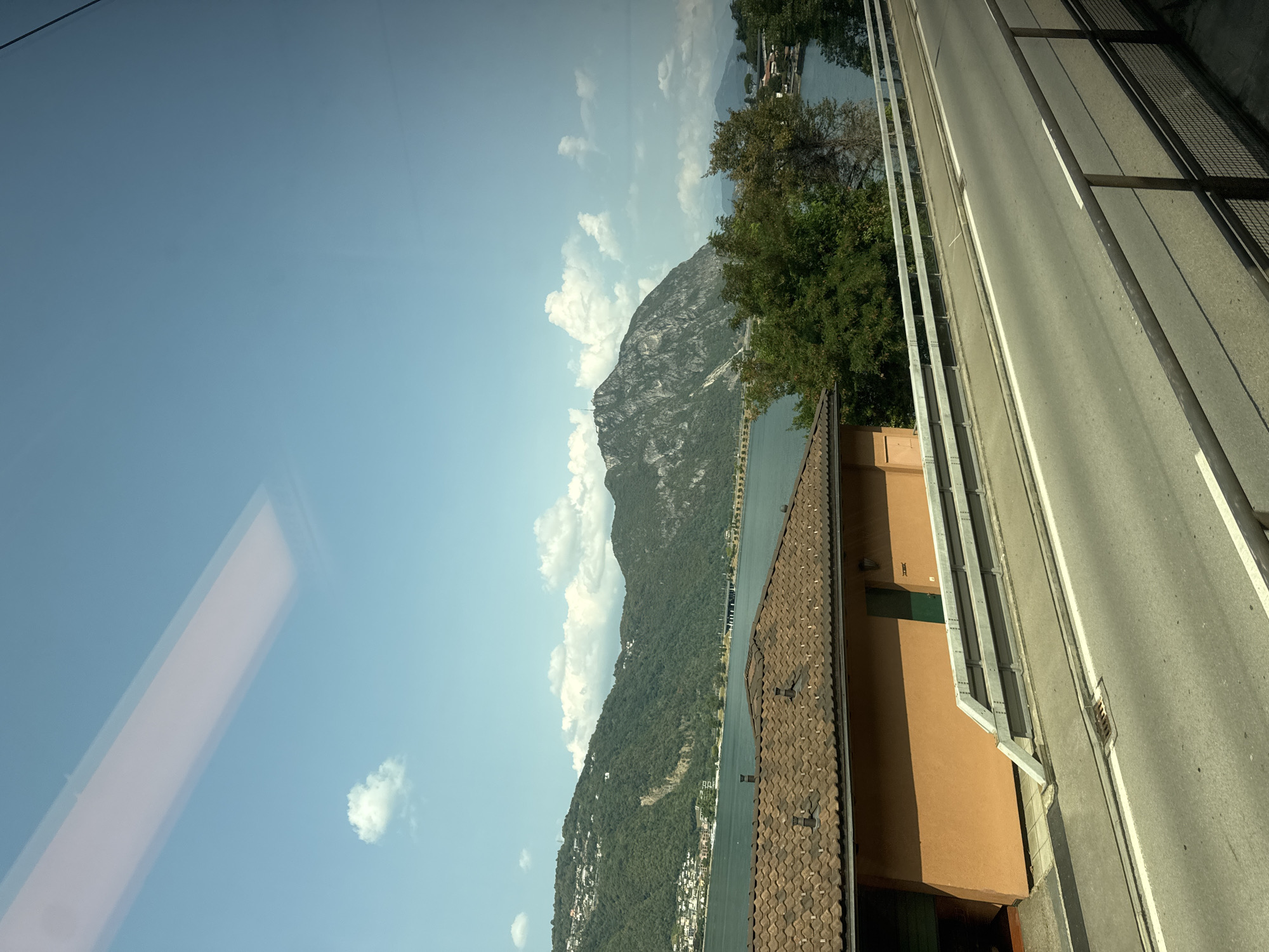 The image captures a scenic view of a lake and mountains, likely from a moving vehicle, as indicated by the reflection on the glass in the upper portion of the frame. In the foreground, a road runs horizontally, bordered by metal railings. To the left of the road is a building with an orange facade and a tiled roof. Beyond the building, a body of water stretches towards a lush, green mountainside dotted with buildings. The sky is blue with scattered white clouds. The location is identified as Biss'one, Switzerland.