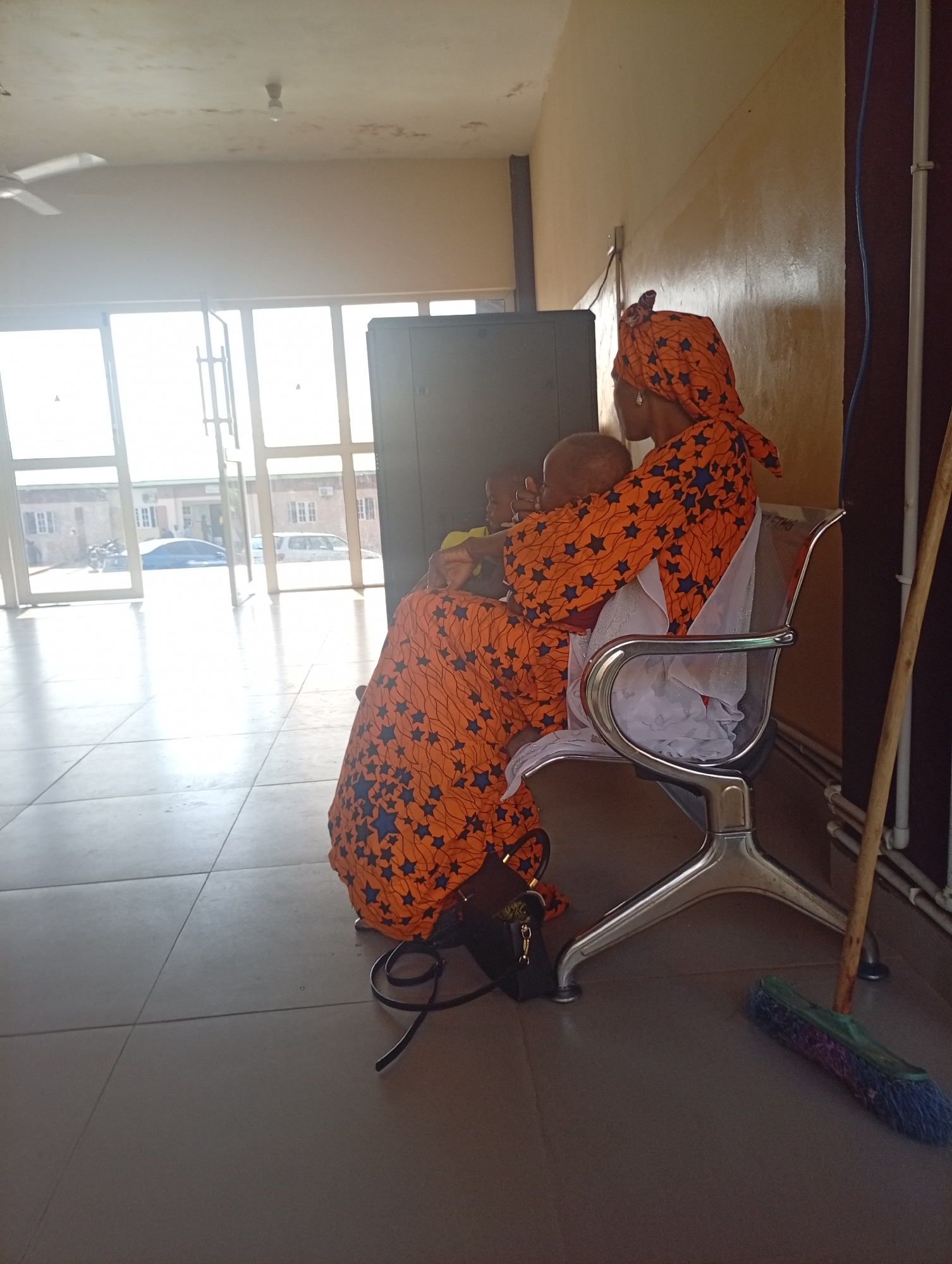 In Gombe, Nigeria, an image captures a woman in an orange and blue star-patterned dress and headscarf sitting in a transparent-seated silver-legged chair. She holds a baby, and another child is partially visible in her lap. The woman's attire suggests a cultural context, possibly a community gathering or public space. The chair is positioned near a large, dark speaker. In the background, large glass doors reveal an outdoor area with buildings and vehicles, indicating a transitional zone between interior and exterior. A broom leans against a wall to the right of the scene, suggesting the space is maintained. The flooring appears to be tiled. The overall impression is of a candid moment within a functional, possibly communal, setting in Gombe.