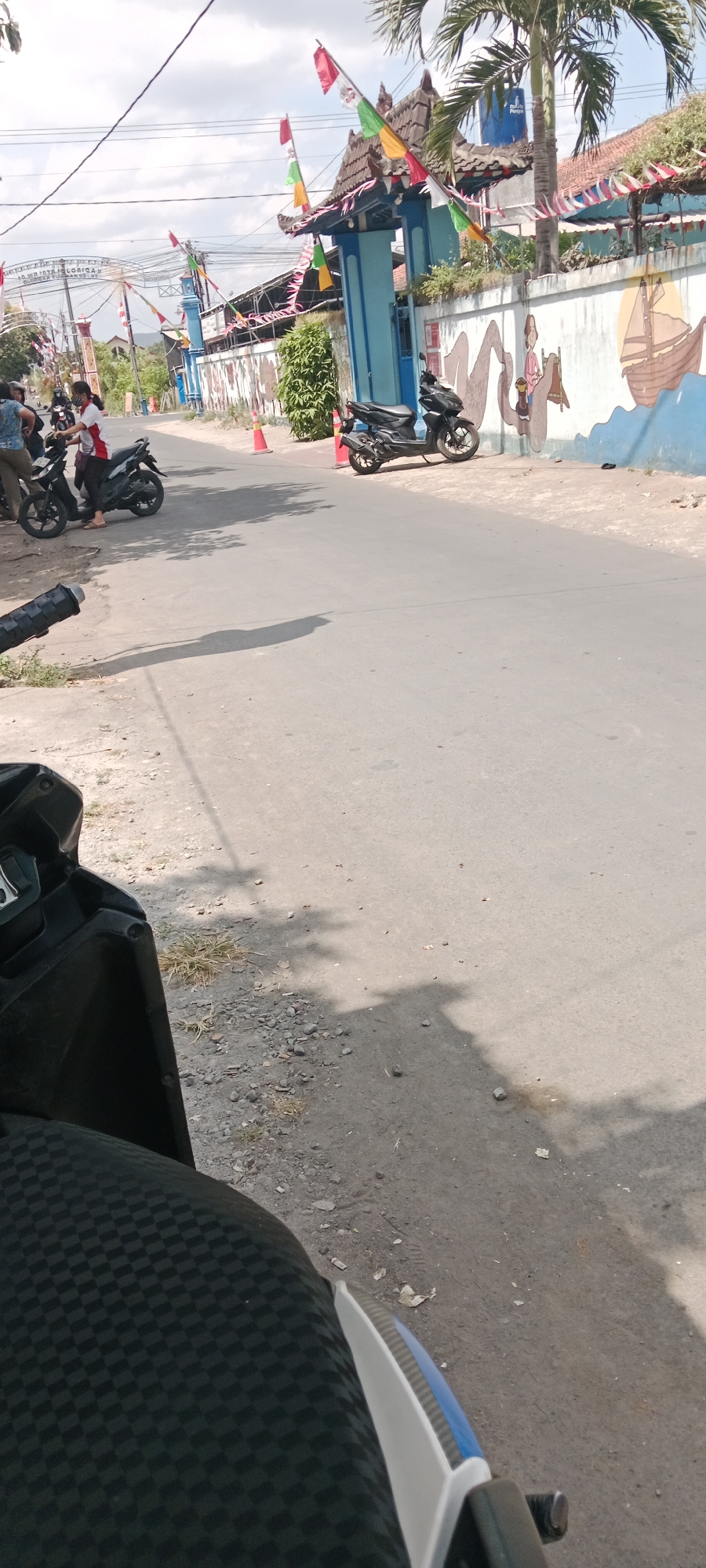 The image shows a street scene in Purwomartani, Indonesia. The perspective is from the seat of a motorcycle, looking forward. To the left, there is a portion of the motorcycle's handlebars and seat visible, featuring a black checkered pattern.
The street itself is paved with asphalt and shows signs of wear and tear, with some gravel and debris scattered along the edges. Sunlight casts shadows of trees and possibly overhead wires onto the road.
On the right side of the street, a building with a blue facade and a traditional roof is visible. The building is decorated with colorful flags and banners, including red and white Indonesian flags. A mural depicting a sailing ship and figures is painted on the exterior wall. Two black motorcycles are parked in front of the building. One is closer to the foreground and appears to be a modern scooter-style motorcycle. Further down the street, another, identical motorcycle is parked.
To the left of the building and street, there are several people gathered around motorcycles. One person in a red and white shirt is standing next to a motorcycle, while others are seated on or near their vehicles. Orange traffic cones are placed along the roadside.
In the background, there are trees and power lines, indicating an urban or semi-urban environment. The sky is partly cloudy, suggesting a bright day. The overall impression is a typical street scene in a residential or commercial area of Purwomartani, possibly during a local celebration or event given the decorations.
