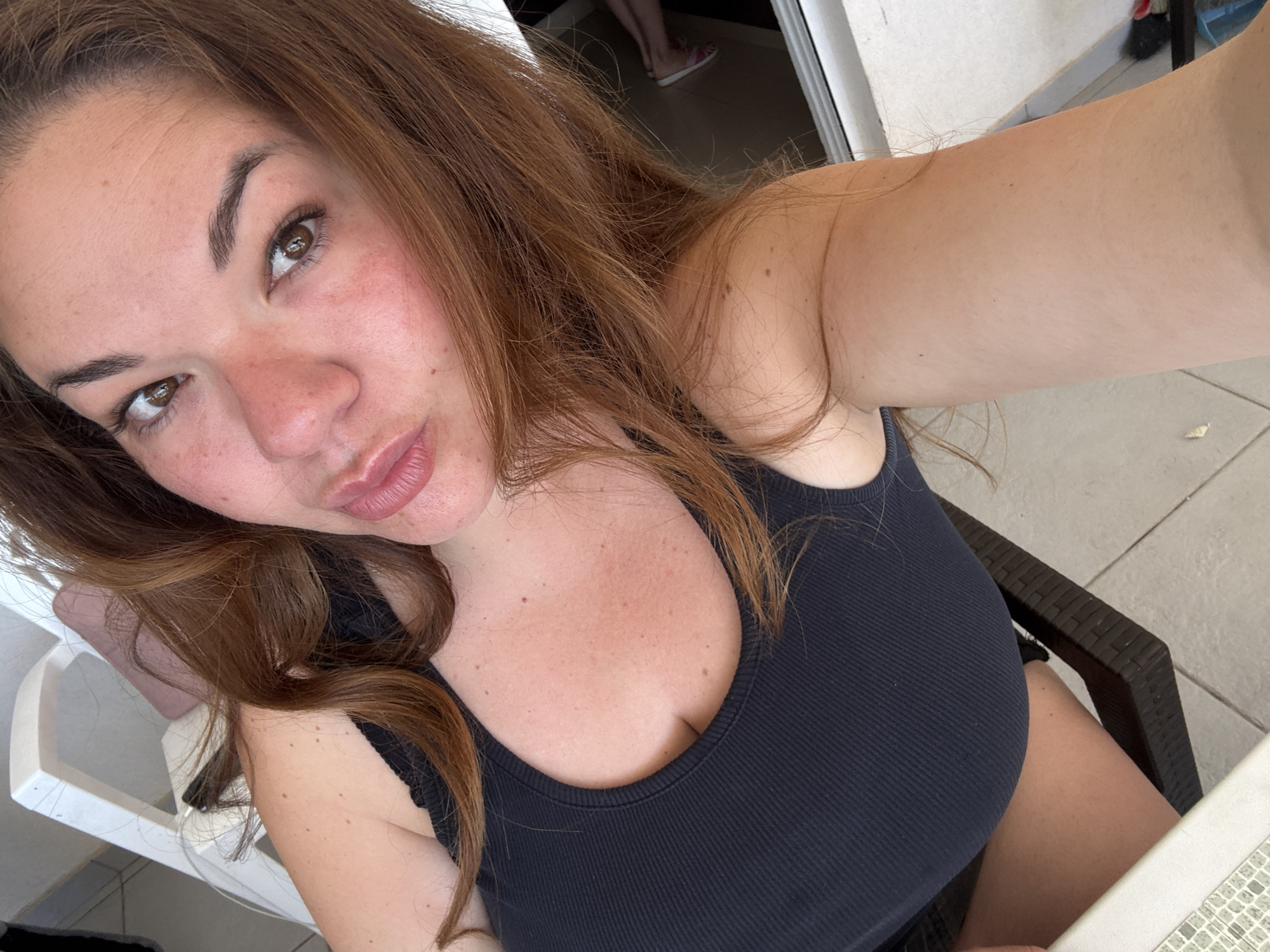 A woman with auburn hair, wearing a black tank top, is taking a selfie. She has light skin with freckles and is looking directly at the camera. The background shows a patio area with white plastic chairs, a dark brown chair, and tile flooring. A doorway is visible in the background with a partial view of another person's legs and feet wearing slip-on shoes. The location may be Bijela, Montenegro.