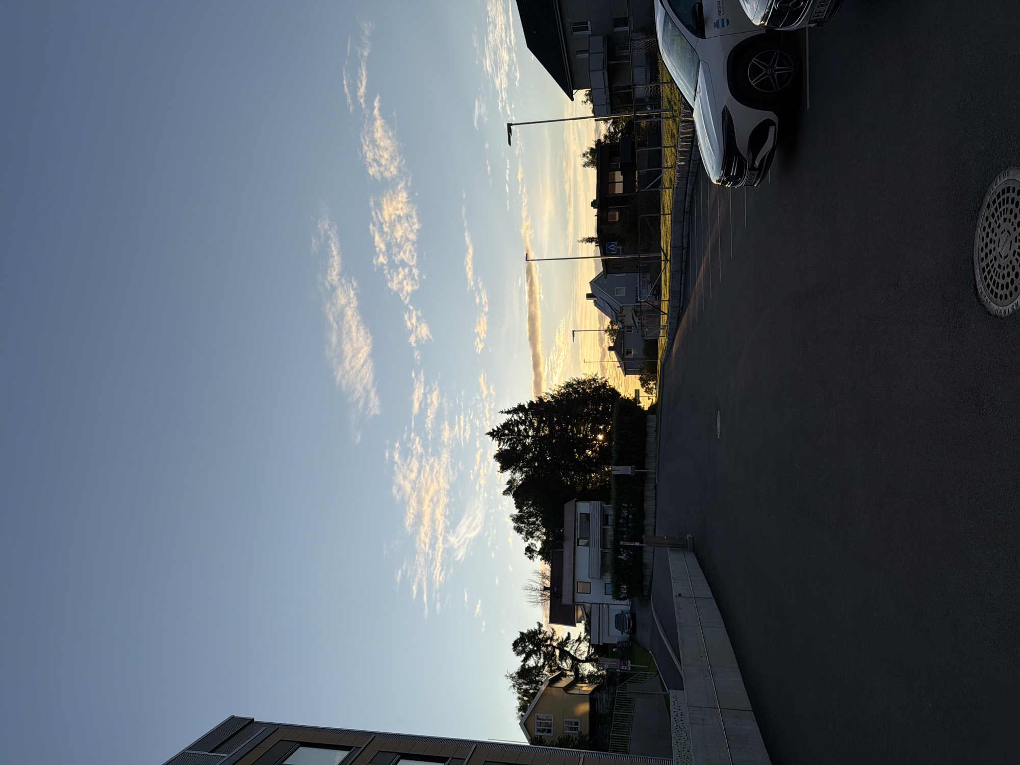 In Harstad, Norway, an asphalt parking lot is bordered by a concrete retaining wall on the left and a street lined with houses and trees on the right. The sky above is a gradient of pale blue to pale orange, with wispy clouds illuminated by the setting sun. A modern white car is parked in the lot, and another car is partially visible to its right. The scene appears to be a residential area, possibly a condominium complex, given the multi-story building on the far left and the architectural style of the houses. The time of day is likely late afternoon or early evening, as indicated by the low angle of the sun and the warm light.