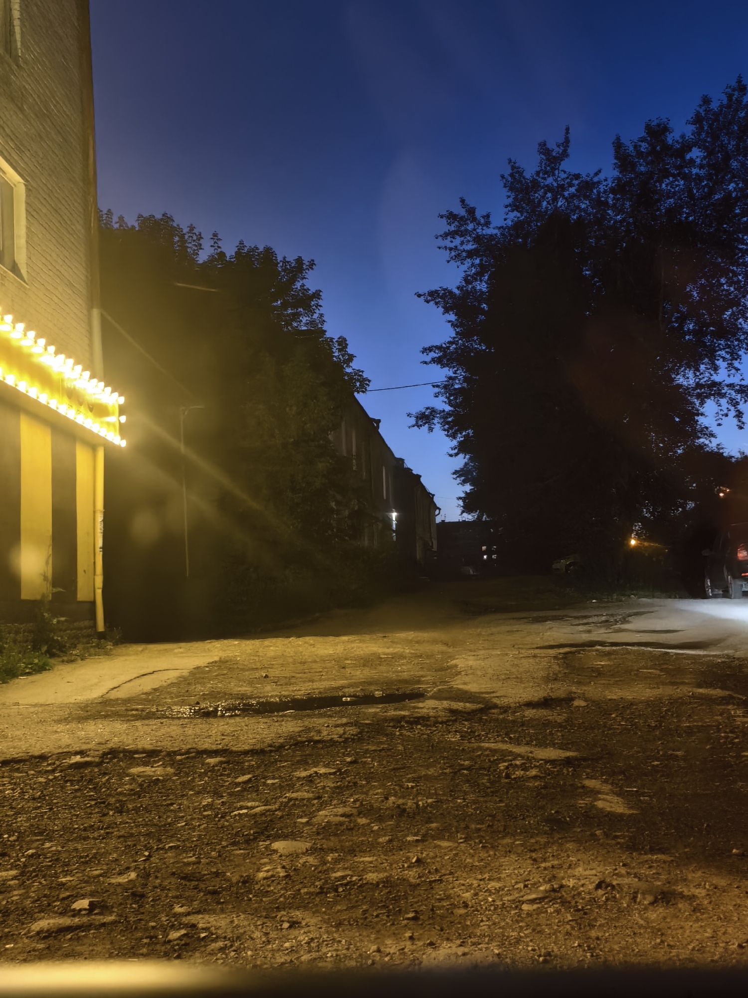 An outdoor night scene in Unknown, Unknown shows a dirt road leading into the distance, flanked by trees and buildings. On the left, a brick building is illuminated by a row of yellow lights, possibly a sign or decorative lighting. The road surface is uneven with visible gravel and puddles, indicating a lack of paving. Further down the road, older buildings with dark windows are visible. Large trees cast shadows across the scene, and the sky transitions from a deep blue to a lighter hue near the horizon. The presence of a vehicle with illuminated taillights on the right suggests a street or parking area. No people are clearly discernible in the image.