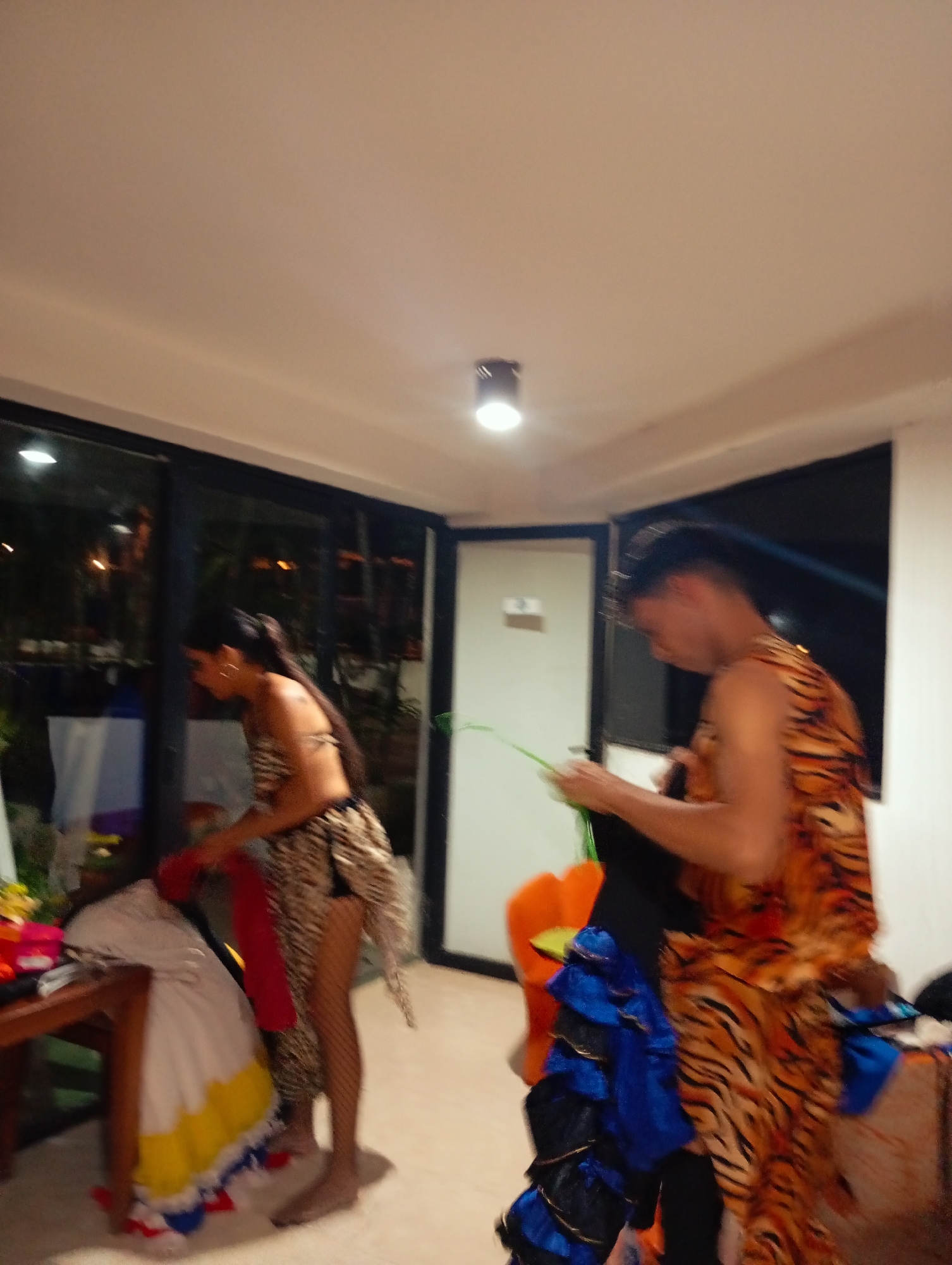 Two people are in a room in Playa El Agua, Venezuela. On the left, a woman in a tiger-striped outfit and fishnet stockings stands near a wooden table with various items on it, including a garment with white, yellow, and blue fabric. She holds a red piece of cloth. On the right, a man wearing a tiger-striped outfit is holding a black garment with blue ruffles. An orange chair is visible in the background. The room has tiled flooring, a black framed sliding glass door on the left, and a white door with a light fixture above in the back. Outside the sliding door, vegetation and lights are visible. A black framed window is on the right.