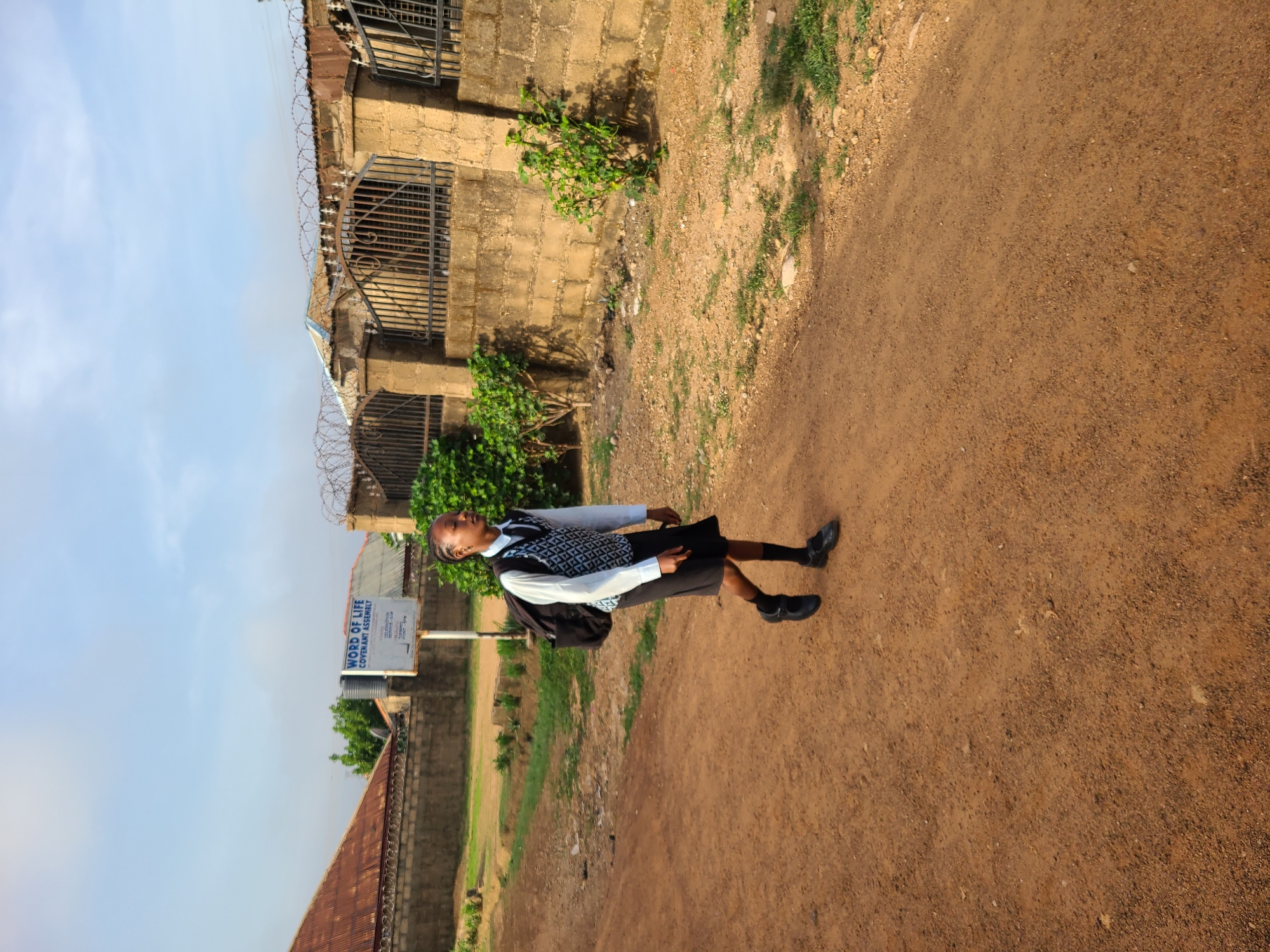 A full shot depicts a student in Barakin Lerawi, Nigeria, walking along a dirt path beside a building and a sign. The student, wearing a white shirt, patterned vest, black skirt, shoes, and socks, also carries a black backpack. The path is reddish-brown dirt. To the left of the path is a weathered building with barred windows topped with barbed wire. Next to it, a sign reads "WORD OF LIFE COVENANT ASSEMBLY." The sky is pale blue with some clouds.