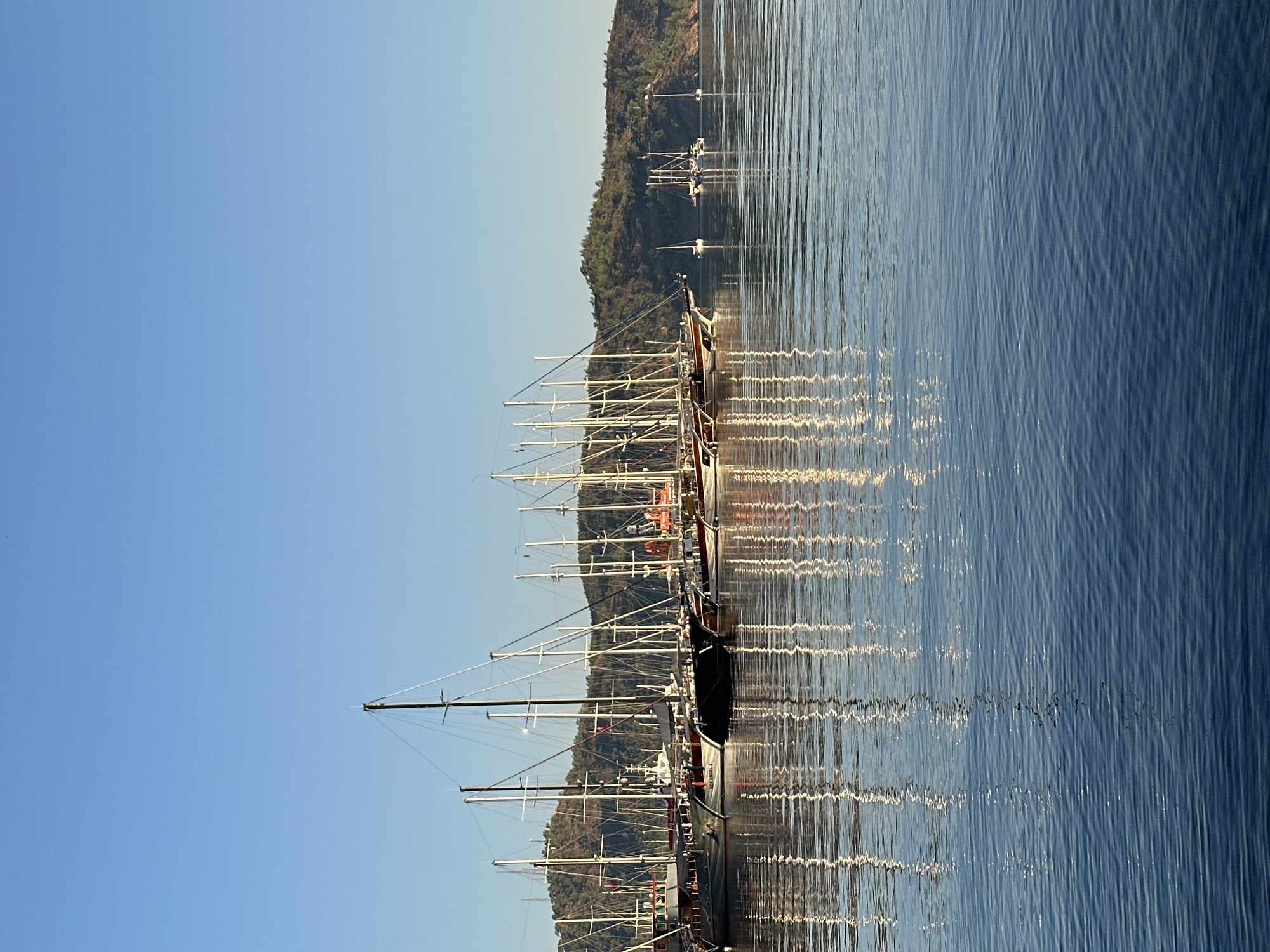 This image depicts a tranquil bay filled with numerous sailboats and gulets docked along a coastline. The boats are lined up close to each other, their masts creating a complex network against the clear blue sky. The calm water reflects the boats and their rigging, creating a symmetrical and serene scene. In the background, a lush, green, tree-covered hillside rises, suggesting a natural, undeveloped landscape bordering the bay. A few smaller boats are visible further out in the water. The overall impression is one of a peaceful harbor, likely in a coastal town or region known for its sailing activities. The clear sky and still water suggest it is either early morning or late afternoon, with soft sunlight illuminating the scene.
This scene is characteristic of the Fethiye region of Turkey, specifically areas like Fethiye Bay or the nearby marina areas where many sailing vessels gather. The combination of the calm, reflective water, the abundance of gulets and sailboats, and the backdrop of verdant hills is a common sight in this part of the Turkish Riviera.